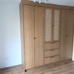 KSD Design - Sylwia Szczerkowska - Jasna, drewniana szafa wnękowa z rattanowymi frontami i złotymi uchwytami, stojąca na podłodze z paneli. Minimalistyczny design, oświetlenie punktowe na suficie.