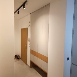 KSD Design - Sylwia Szczerkowska - Nowoczesna szafa wnękowa z szarymi frontami i drewnianymi akcentami, wkomponowana w przedpokój z oświetleniem szynowym i drewnianymi drzwiami wejściowymi.