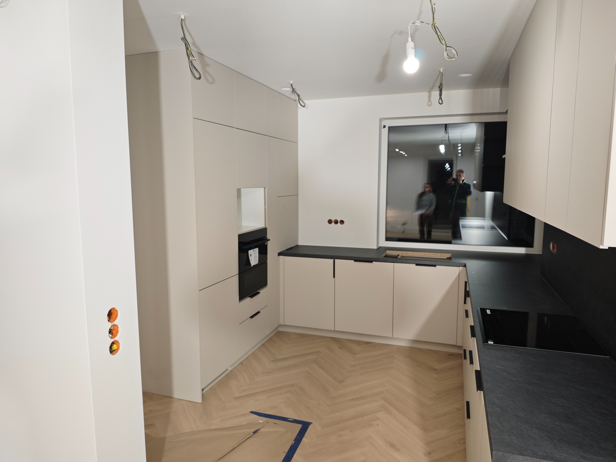 Jasna kuchnia z czarnym blatem i sprzętem AGD w zabudowie. Na podłodze parkiet ułożony w jodełkę. Widoczne niedokończone prace elektryczne i zabezpieczenia podłogi.