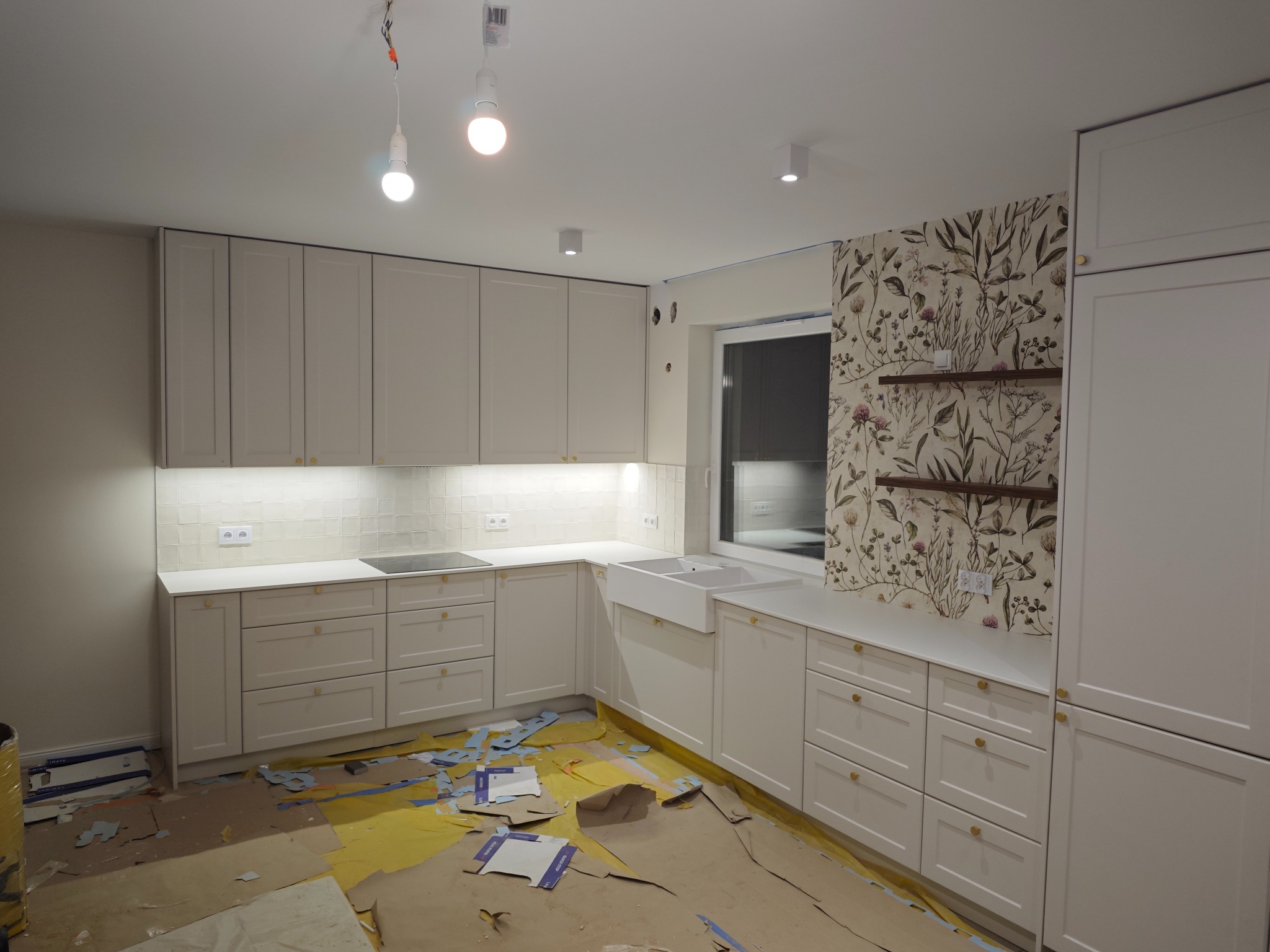 Jasna kuchnia z białymi frontami i złotymi uchwytami, zlew farmerski, tapeta w kwiaty. Na podłodze resztki folii zabezpieczającej, co sugeruje świeży montaż.