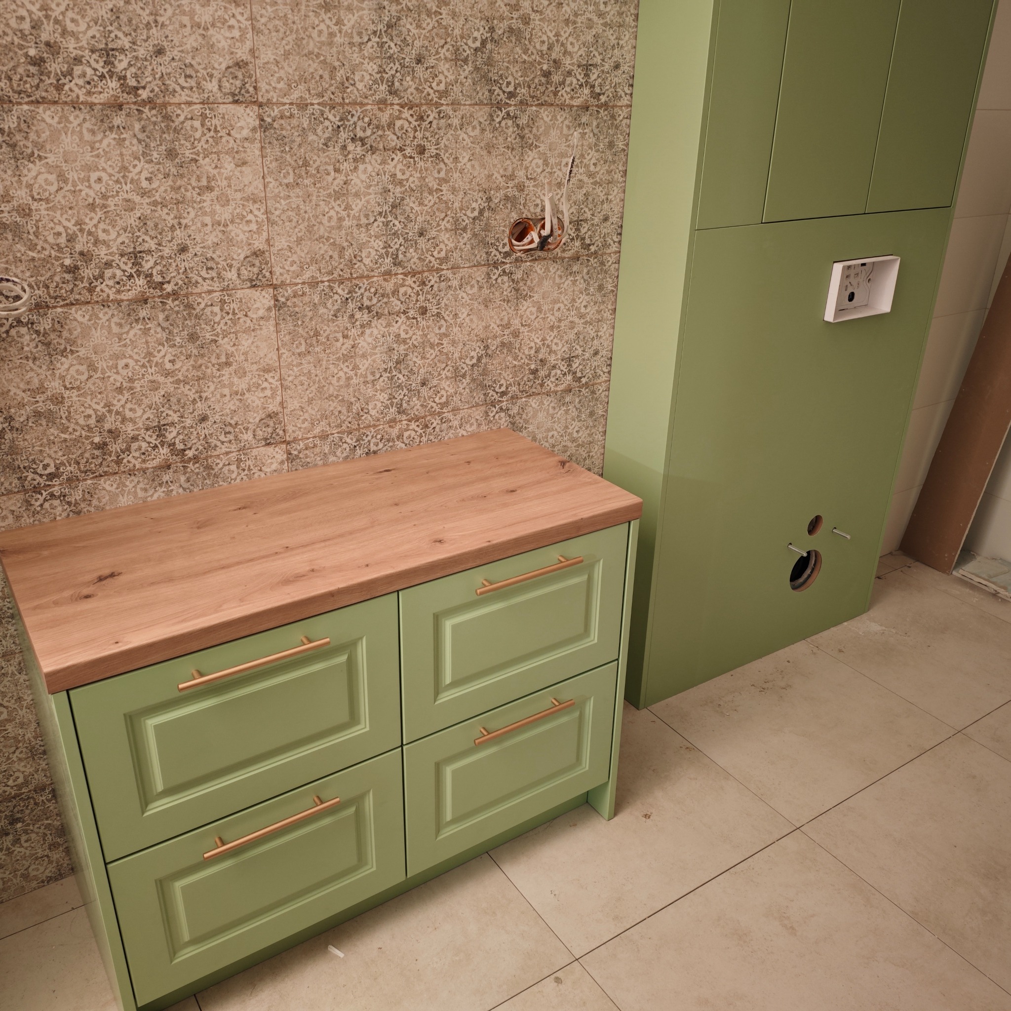 Zielona szafka łazienkowa z drewnianym blatem i złotymi uchwytami na tle wzorzystych płytek i zabudowy geberit. Widoczne przyłącza hydrauliczne.