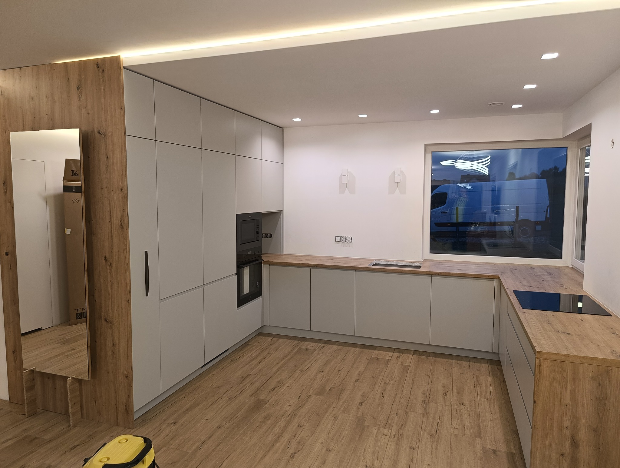 Nowoczesna kuchnia na wymiar z szarymi frontami i drewnianymi blatami. Widoczne zabudowane sprzęty AGD i duże okno z odbijającym się samochodem dostawczym.