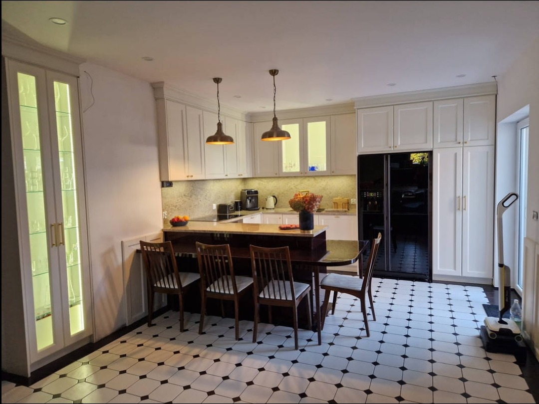 Jasna kuchnia z wyspą i czarną lodówką. Białe szafki z oświetleniem LED i złotymi uchwytami. Podłoga w czarno-białą szachownicę. Dwie lampy wiszące nad wyspą.