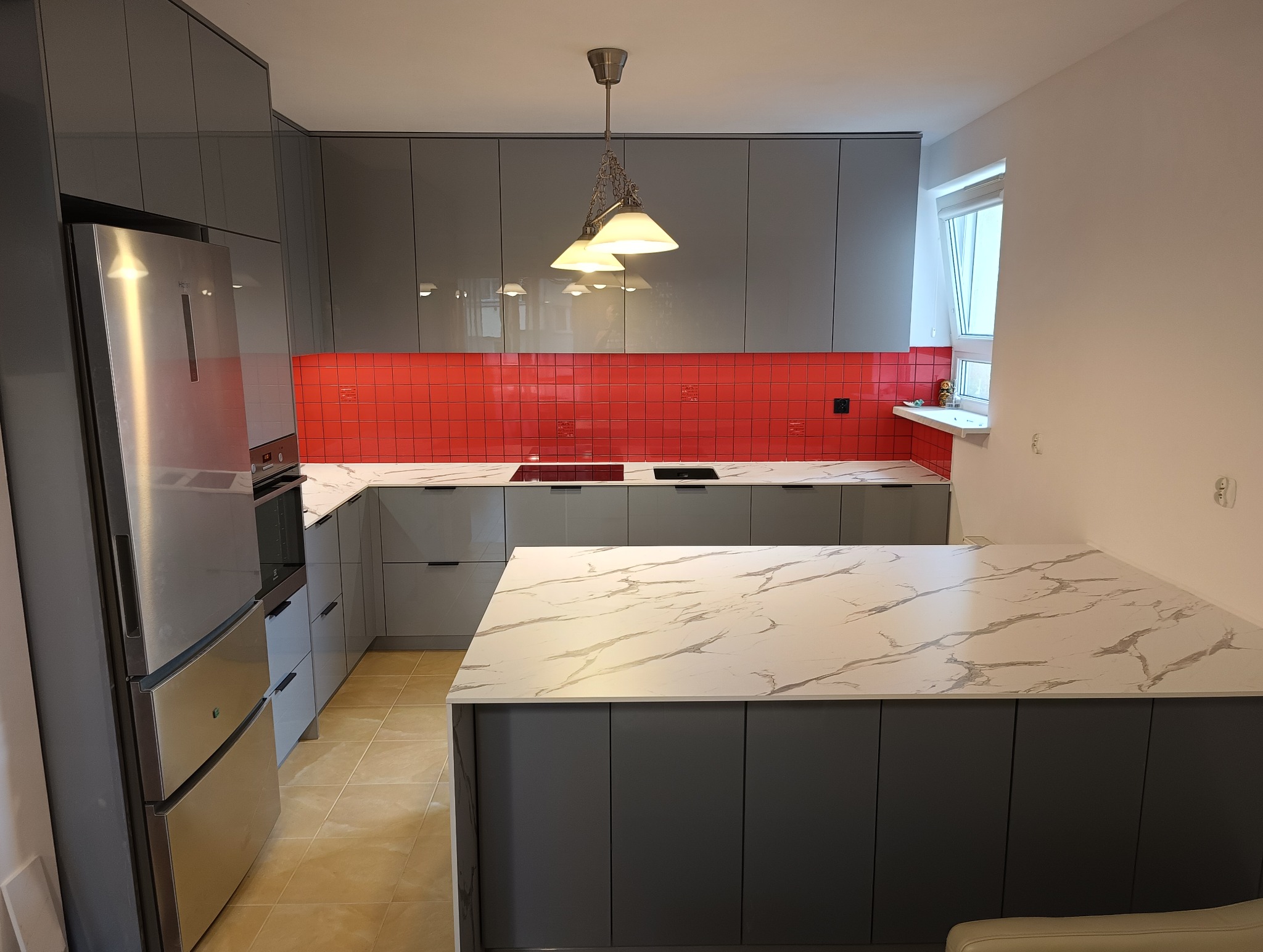 Szara kuchnia z wyspą i czerwonym panelem nad blatem. Nowoczesne szafki bez uchwytów, marmurowy blat. Oświetlenie punktowe.