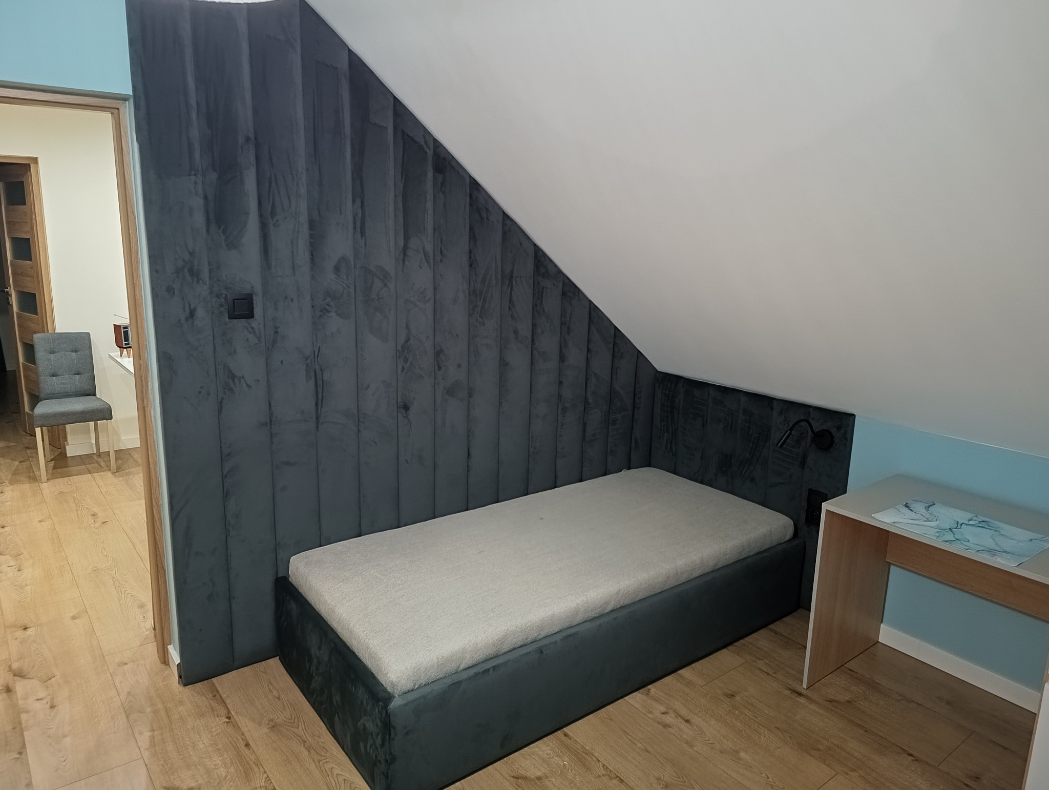 Tapicerowane panele ścienne za łóżkiem w pokoju na poddaszu w Kalwarii Zebrzydowskiej. Ciemnoszary welur, pionowe pasy, lampka nocna. Nowoczesny design.