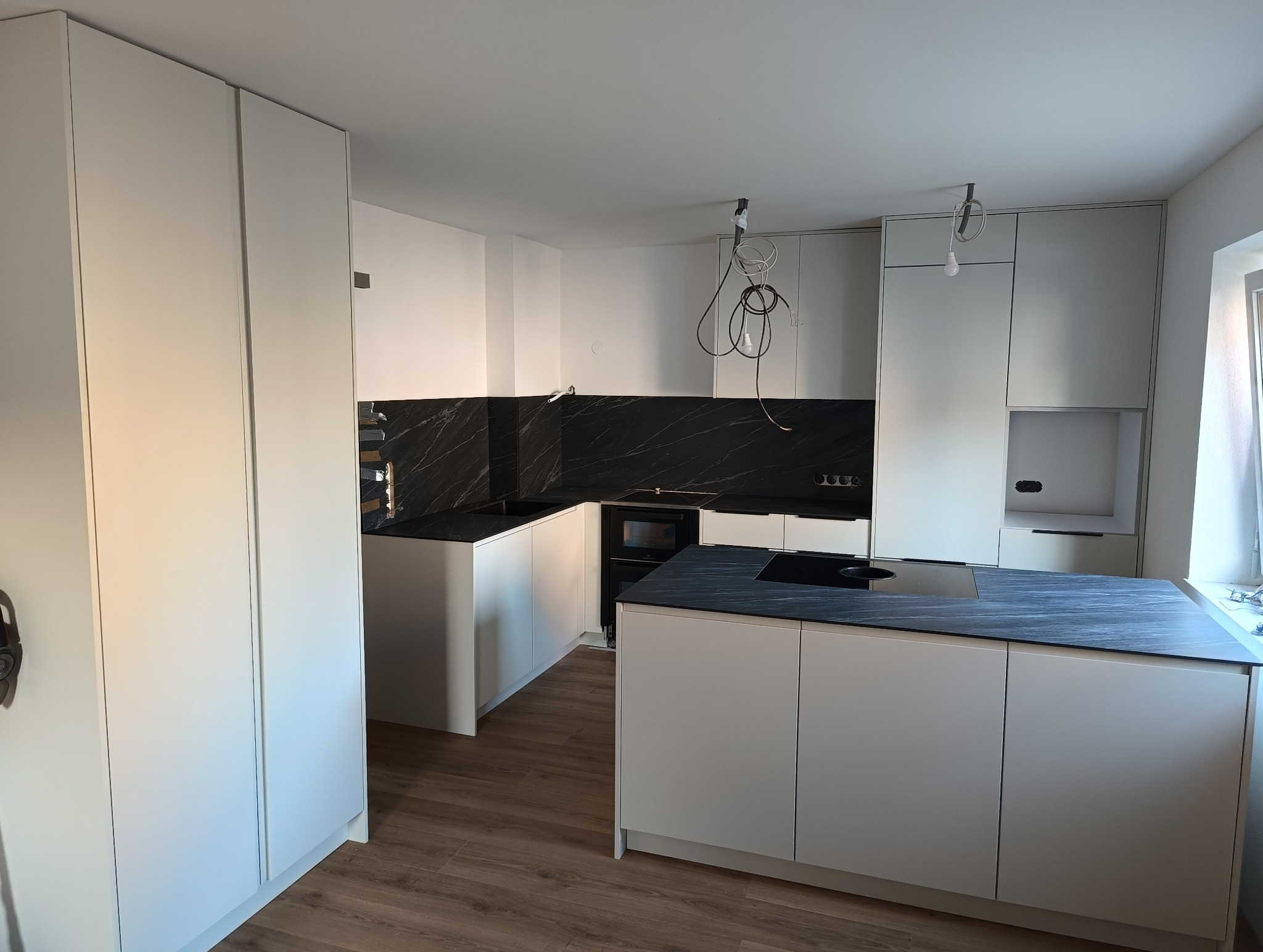 Nowoczesna kuchnia z białymi frontami i czarnym blatem imitującym marmur. Wyspa kuchenna z płytą indukcyjną. Widoczne wiszące kable od oświetlenia.