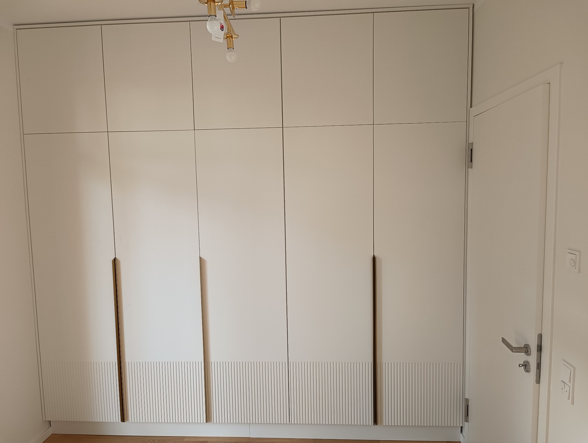 Biała szafa wnękowa z frezowanymi frontami i długimi, smukłymi uchwytami w odcieniu złota, dopasowana do wysokości pomieszczenia, widok na fragment drzwi.