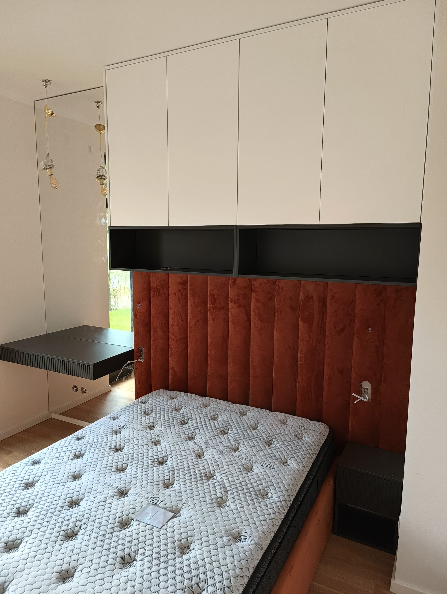 Nowoczesna sypialnia z łóżkiem z wezgłowiem z brązowego weluru, szafkami wiszącymi i biurkiem. Minimalistyczny design, stonowana kolorystyka, oświetlenie punktowe.