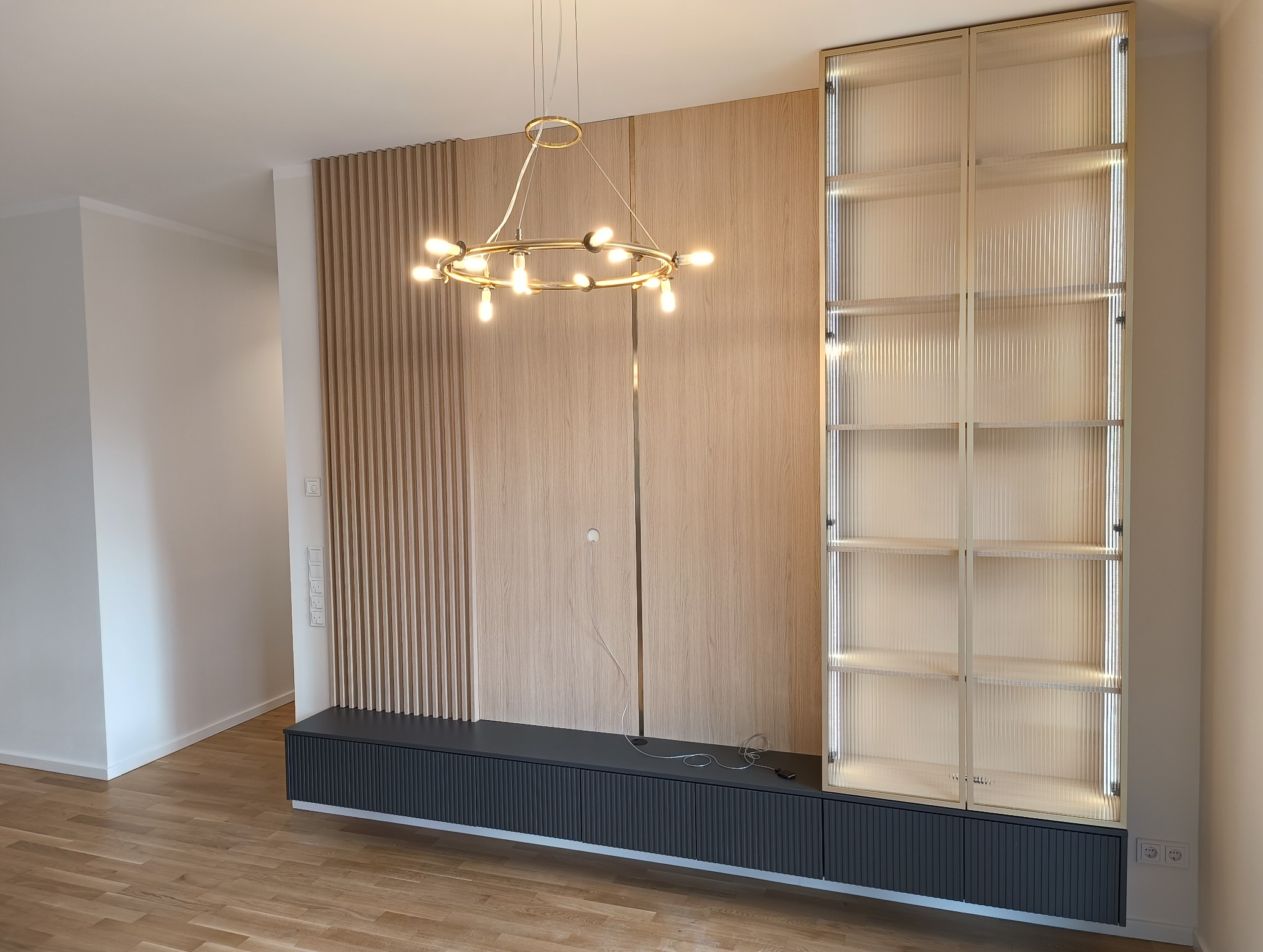 Nowoczesna zabudowa ściany z lamelami, szafką RTV i przeszkloną witryną z podświetleniem LED. Złoty żyrandol dodaje elegancji. Minimalistyczny design.