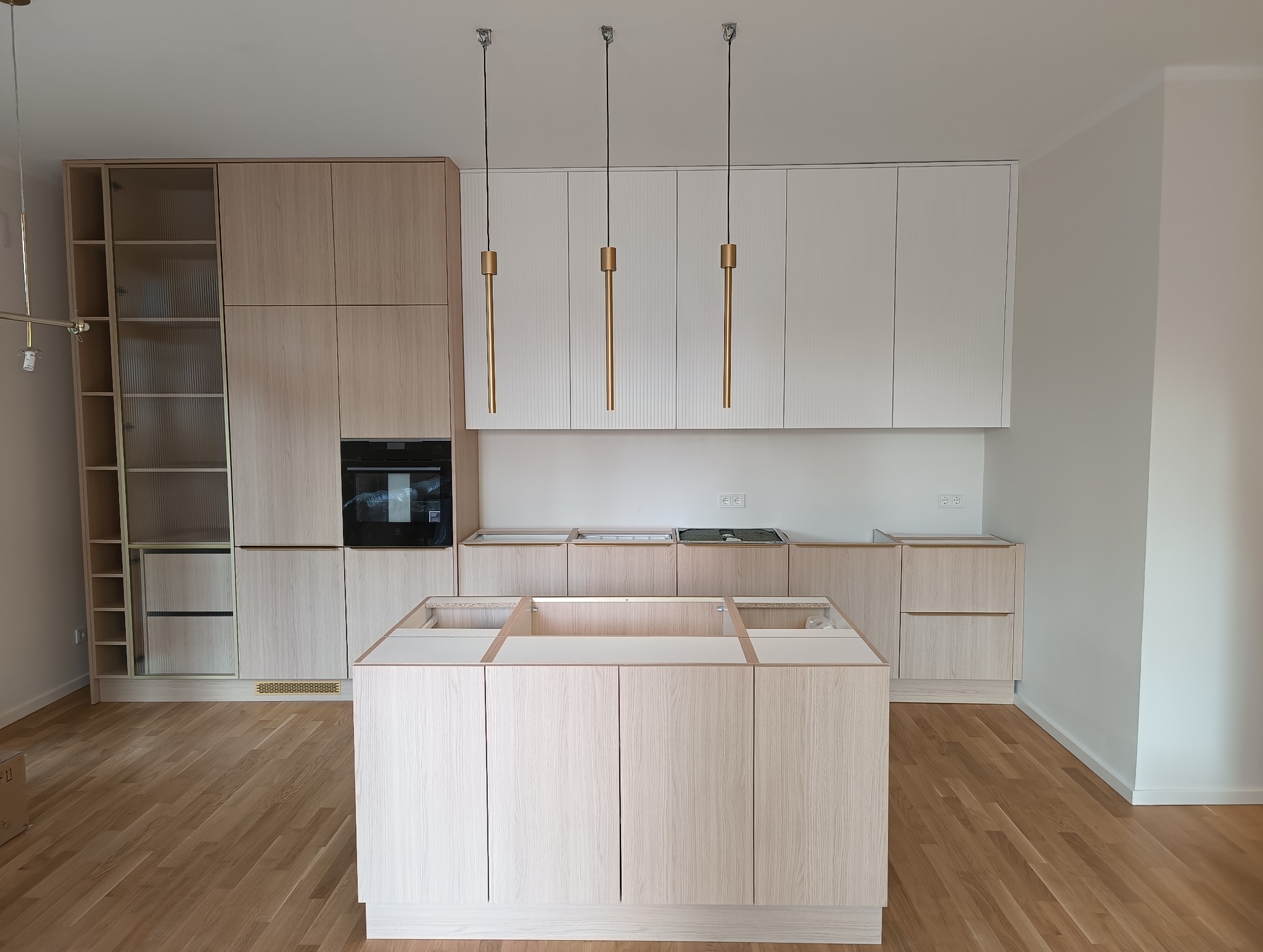 Jasna, nowoczesna kuchnia z wyspą w trakcie montażu. Widoczne szafki z drewnianym fornirem i białe górne szafki. Nad wyspą wiszą trzy złote lampy.