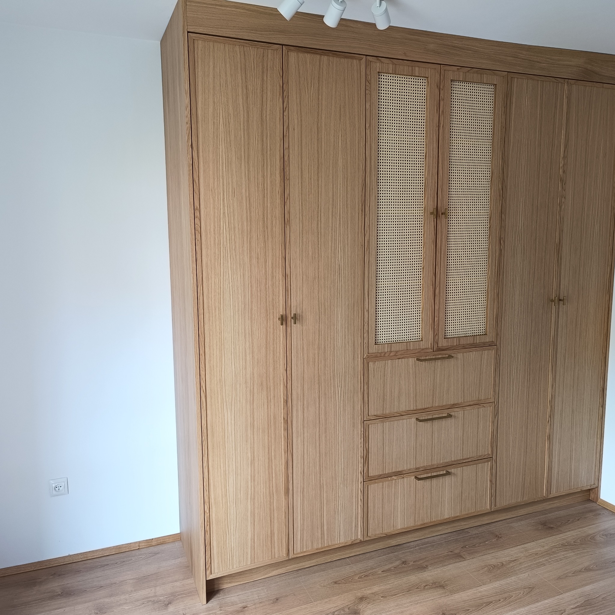 Jasna, drewniana szafa wnękowa z rattanowymi frontami i złotymi uchwytami, stojąca na podłodze z paneli. Minimalistyczny design, oświetlenie punktowe na suficie.