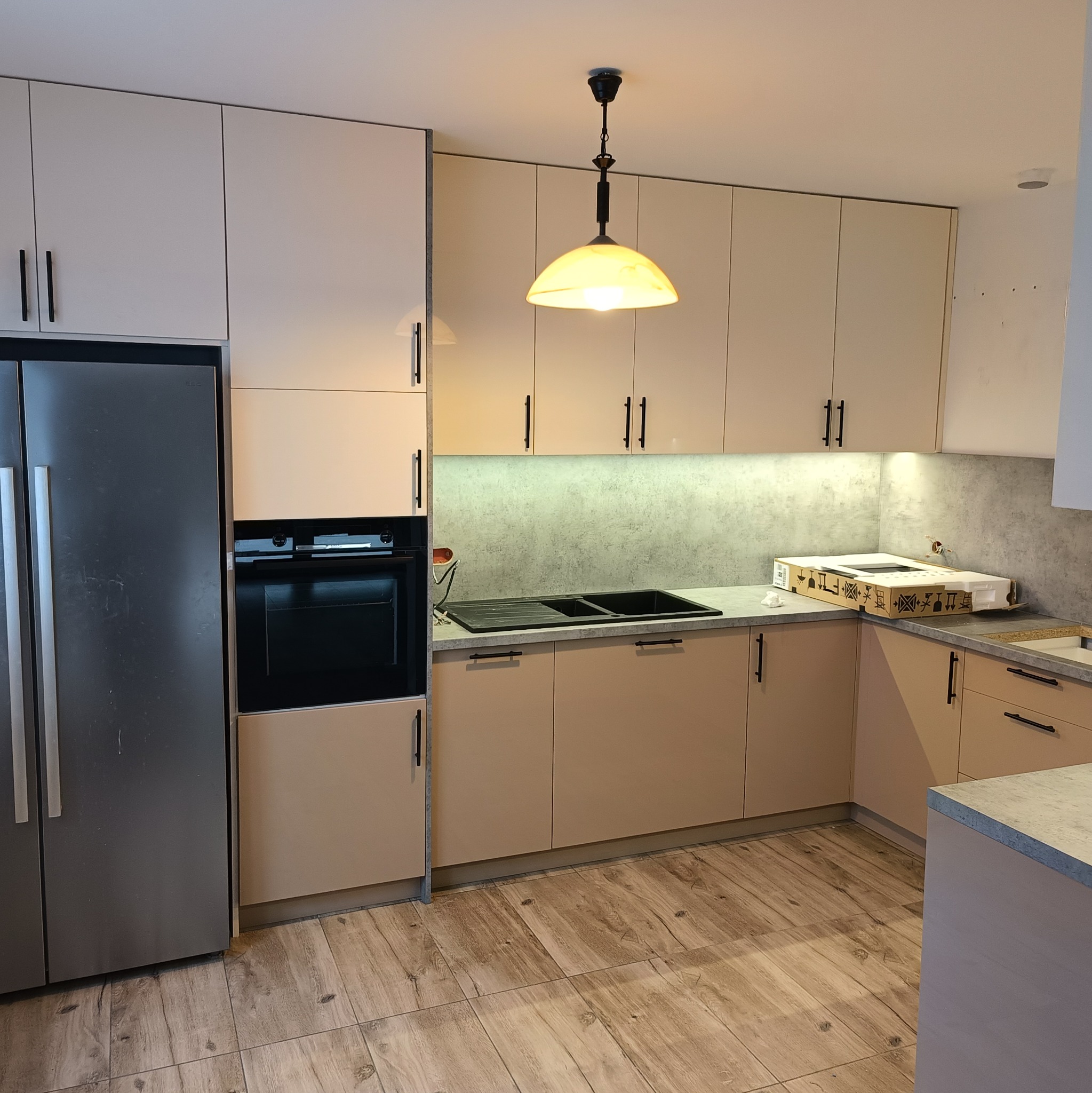 Jasna kuchnia na wymiar z beżowymi frontami i czarnymi uchwytami. Widoczny zlew, piekarnik i lodówka. Na blacie karton z nowym sprzętem. Drewnopodobna podłoga.