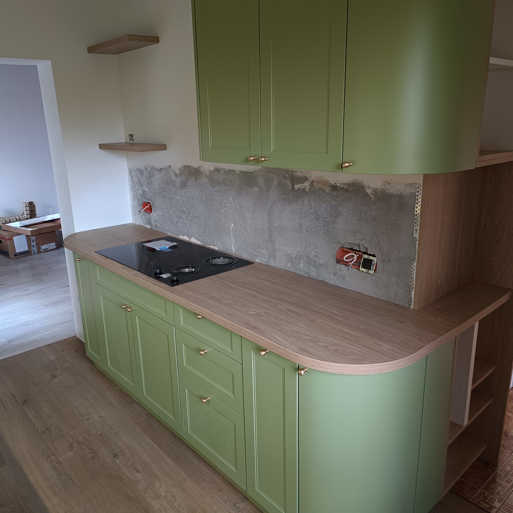 Kremowo-zielona zabudowa kuchenna z drewnianym blatem i złotymi uchwytami, płyta indukcyjna. Widoczne niedokończone ściany i instalacja elektryczna. Ujęcie z perspektywy.