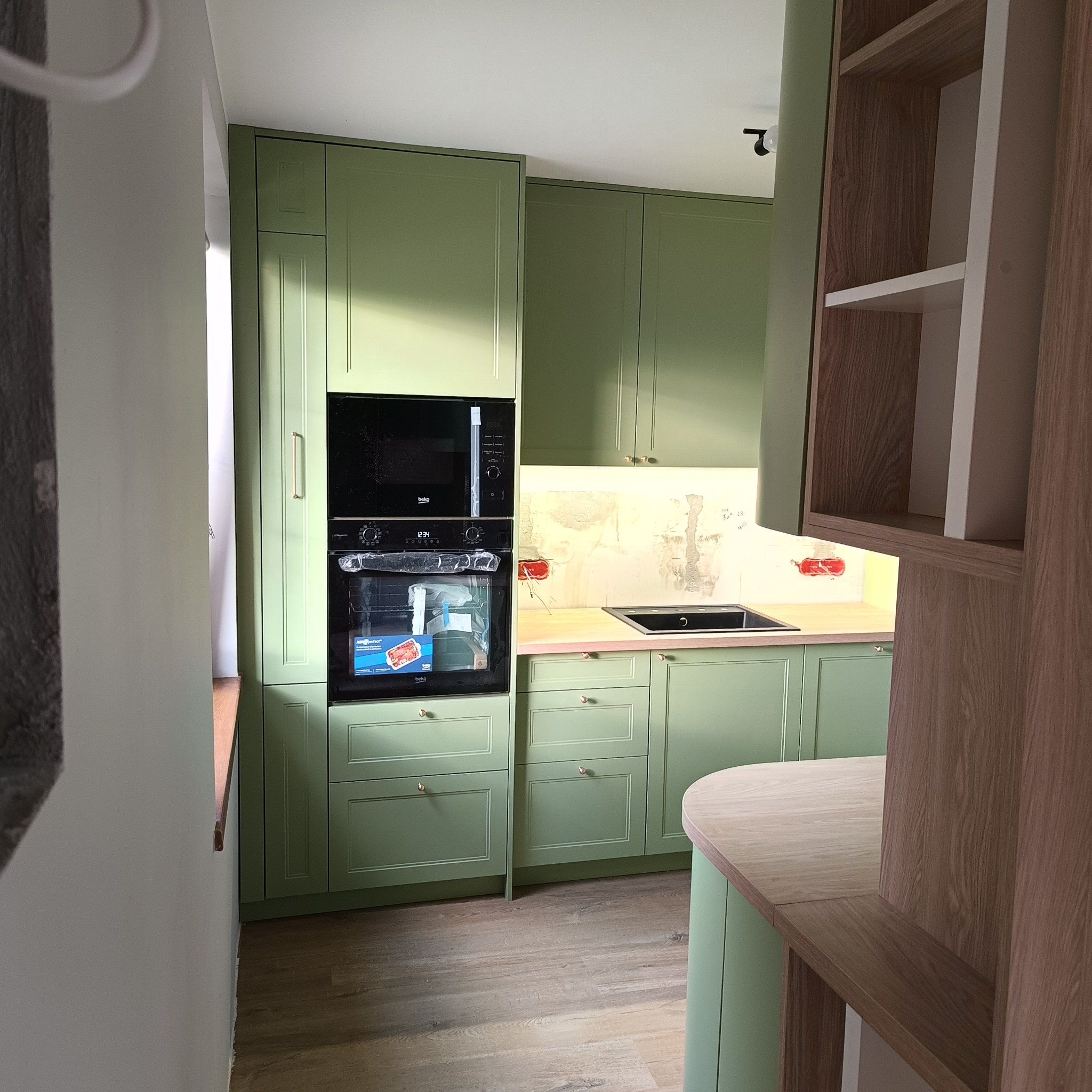Jasnozielona zabudowa kuchenna z drewnianym blatem i sprzętami AGD. Widoczne szafki górne i dolne, piekarnik oraz płyta grzewcza. Styl klasyczny z nowoczesnymi akcentami.