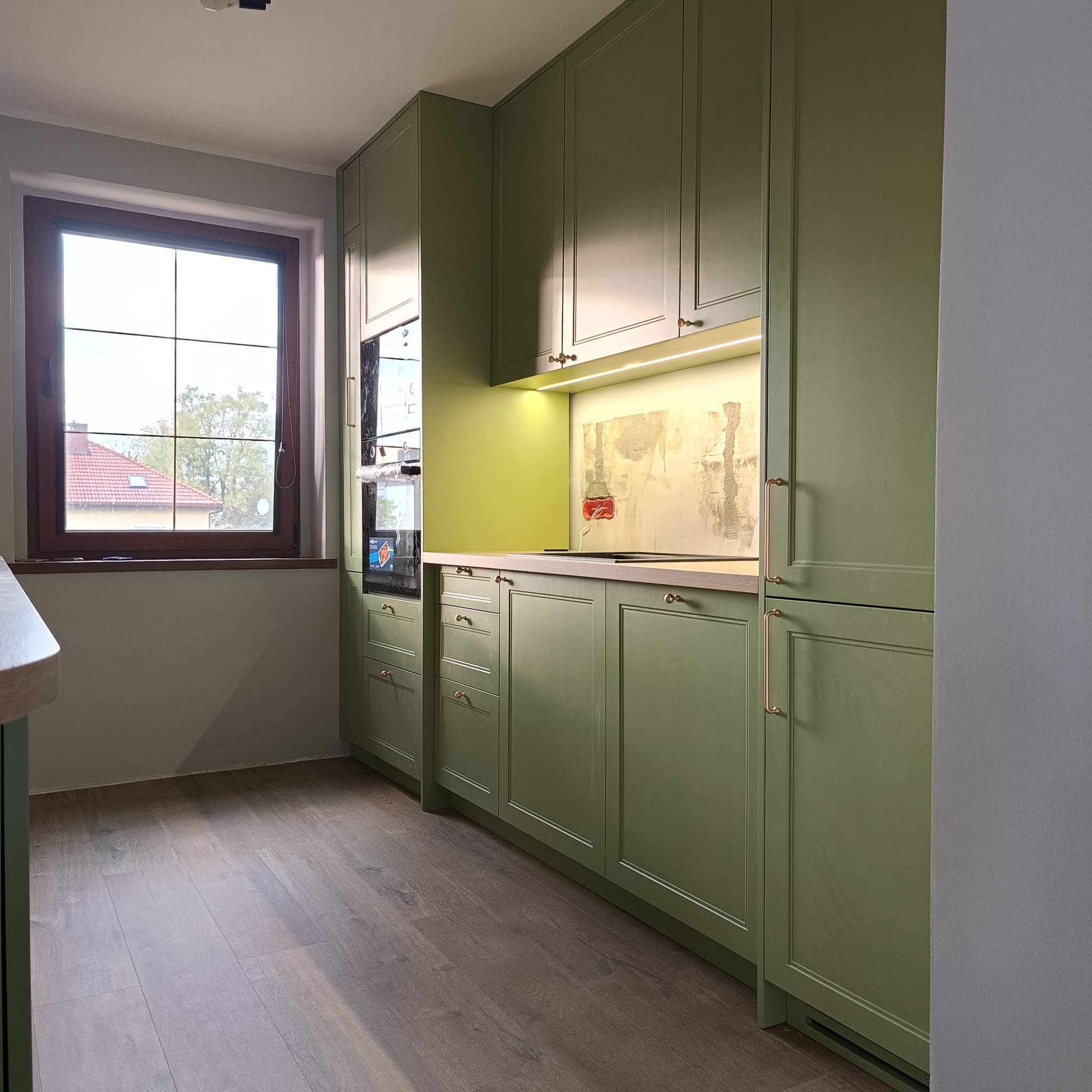 Nowoczesna kuchnia w odcieniach zieleni z drewnianymi akcentami i oświetleniem LED. Widoczne okno z widokiem na dom z czerwoną dachówką. Eleganckie wykończenie.