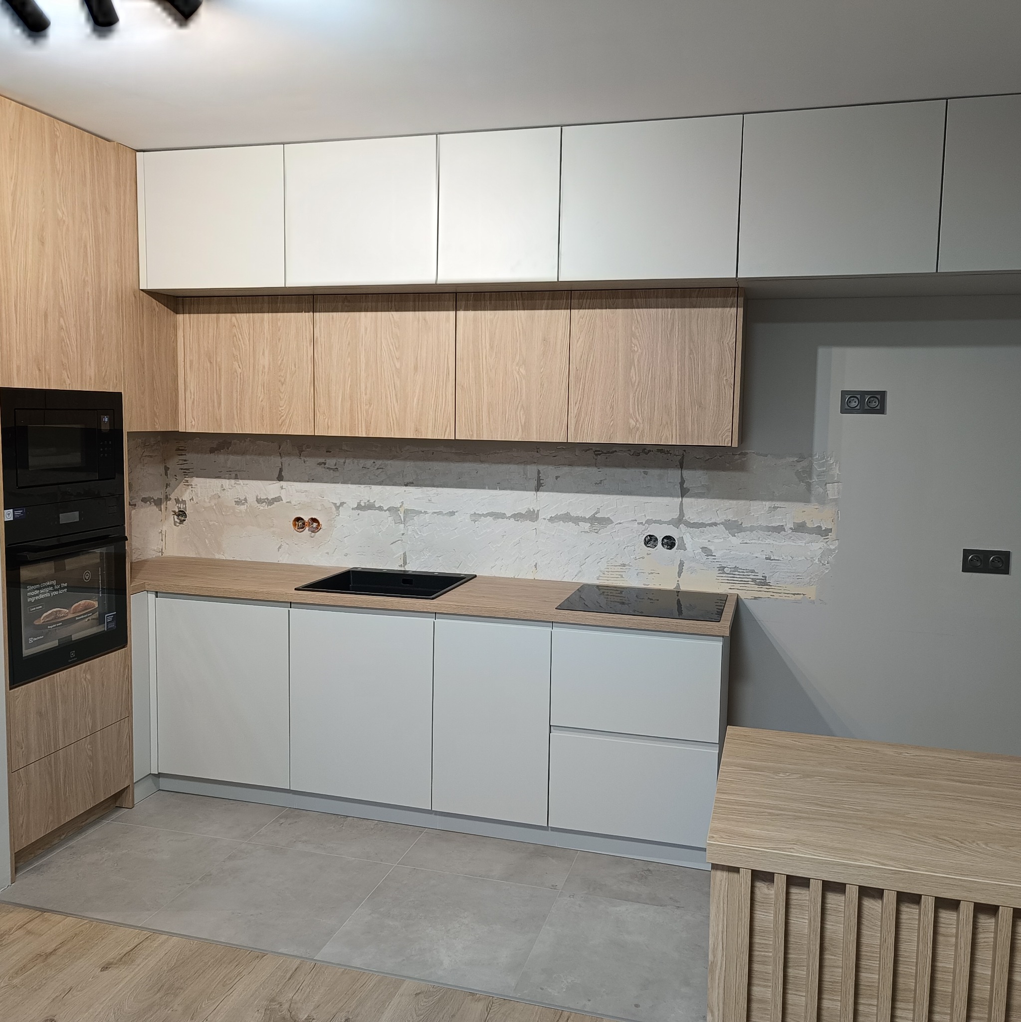 Jasna kuchnia z drewnianymi i białymi frontami szafek, czarną płytą indukcyjną i piekarnikiem w zabudowie. Ściana nad blatem częściowo wykończona, widoczne gniazdka.