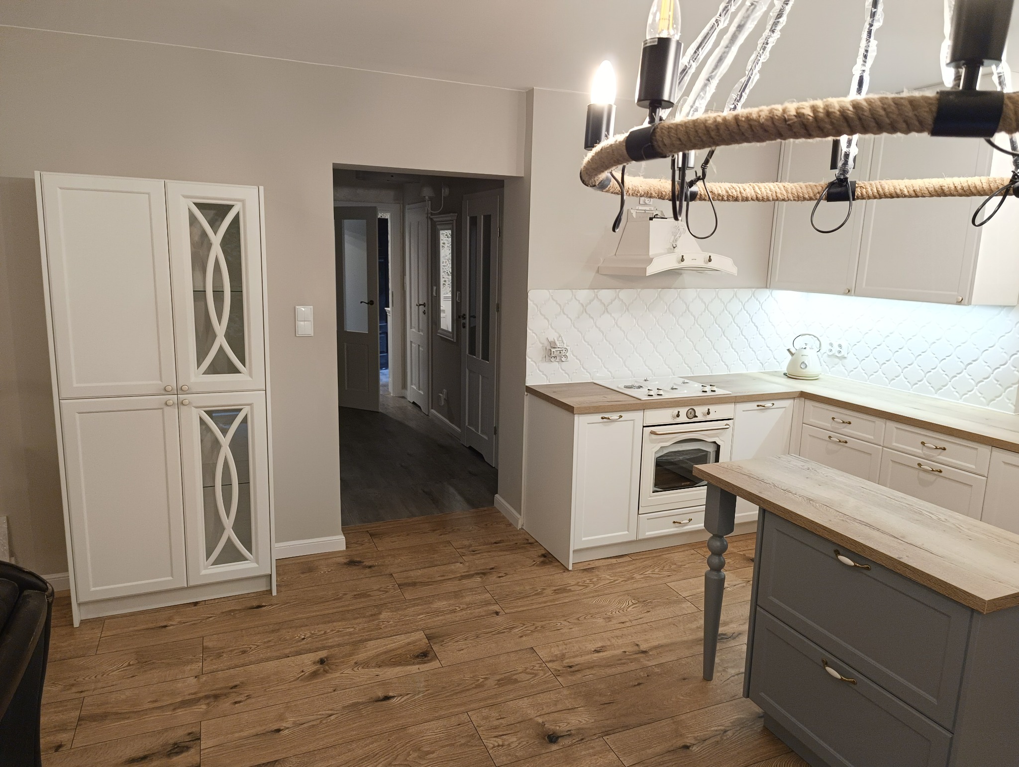 Jasna kuchnia z drewnianą podłogą i wyspą. Białe szafki z frezowanymi frontami, dekoracyjny żyrandol z liny. Widok na dalsze pomieszczenia.