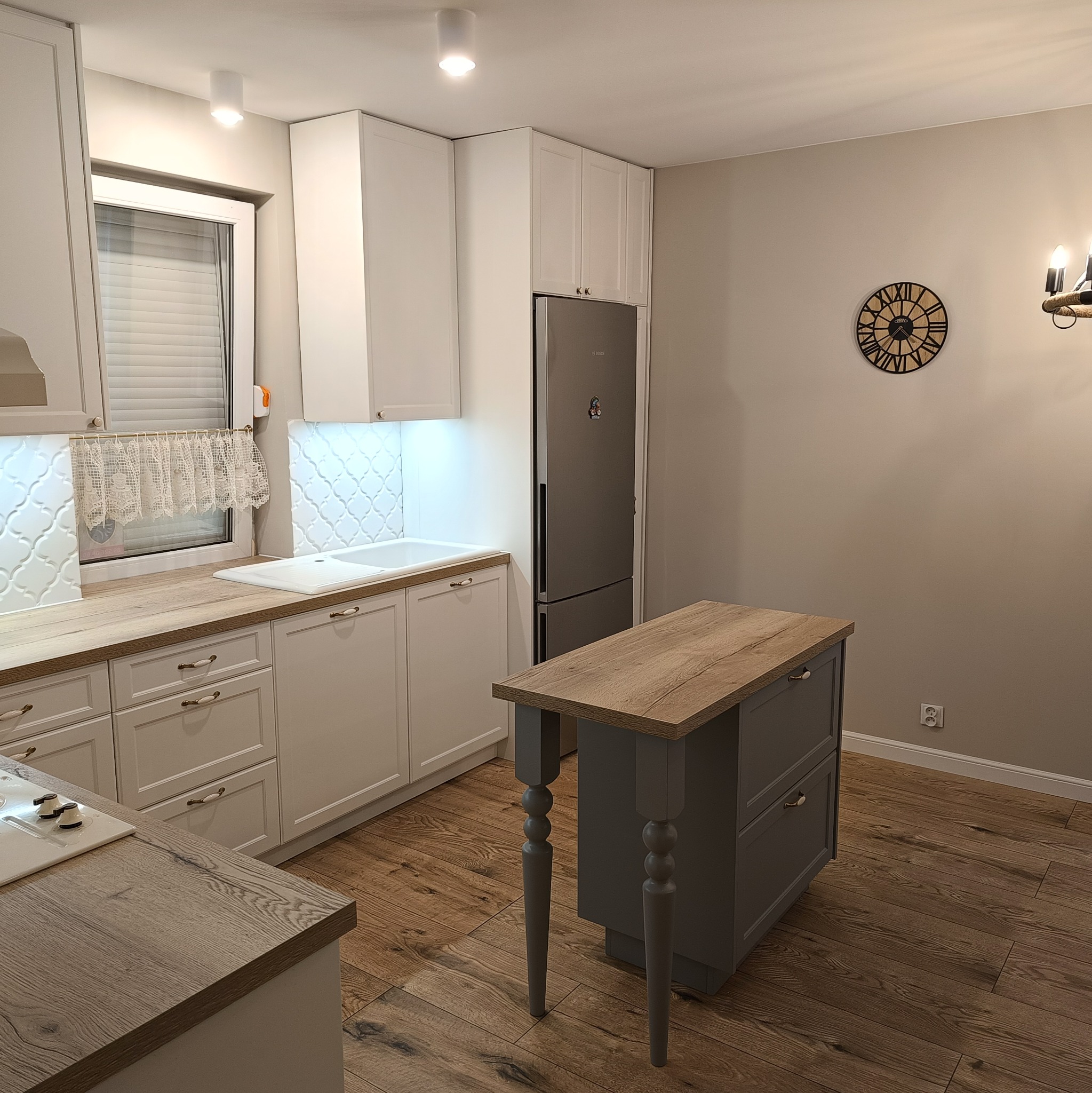 Jasna kuchnia z wyspą na drewnianej podłodze. Białe szafki z frezowanymi frontami, drewniany blat i szara wyspa z toczonymi nogami. Na ścianie dekoracyjny zegar.