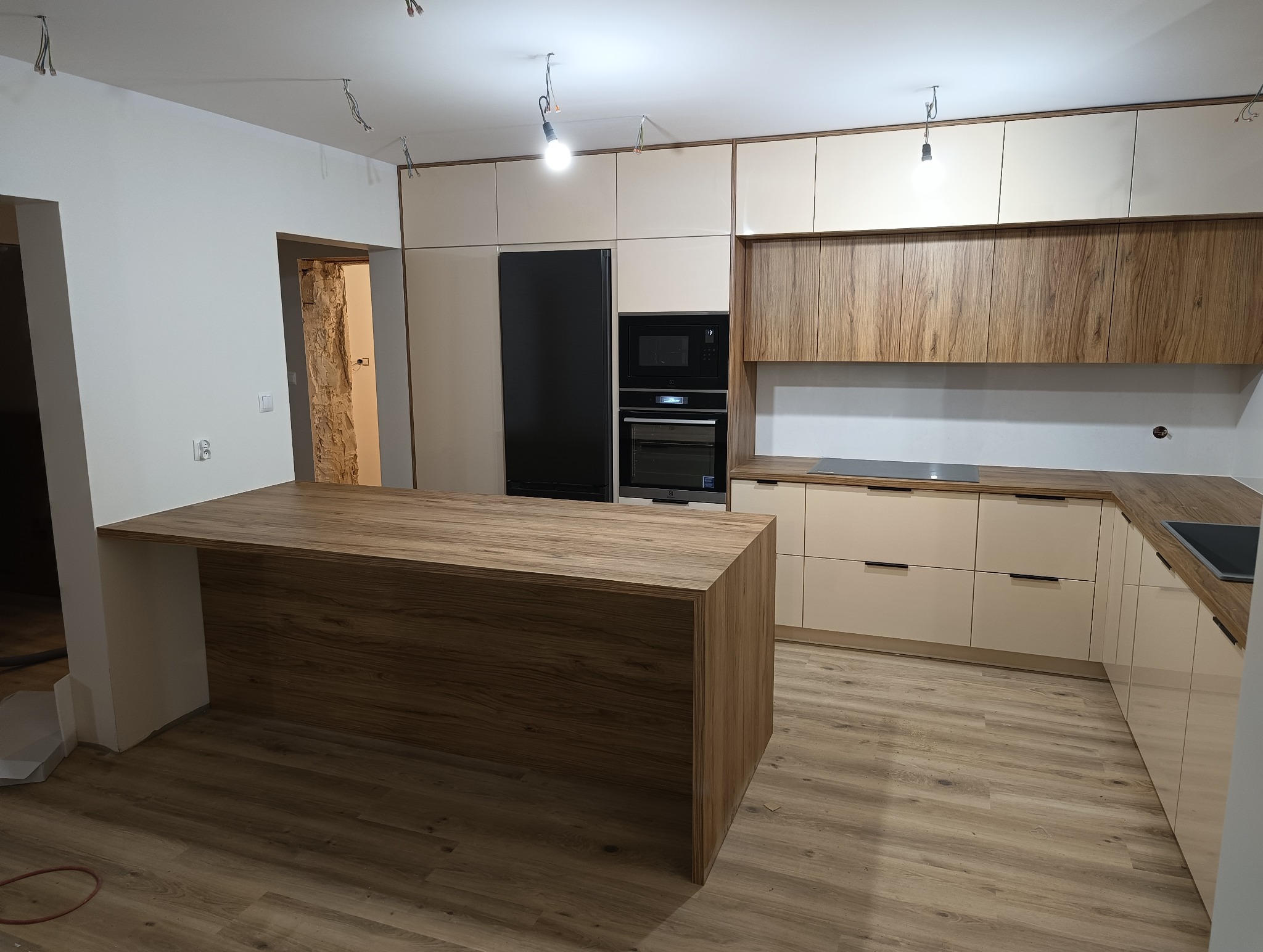 Nowoczesna kuchnia na wymiar z wyspą, fronty w odcieniach beżu i drewna, czarny sprzęt AGD w zabudowie. Widoczne niedokończone wykończenia sufitu.