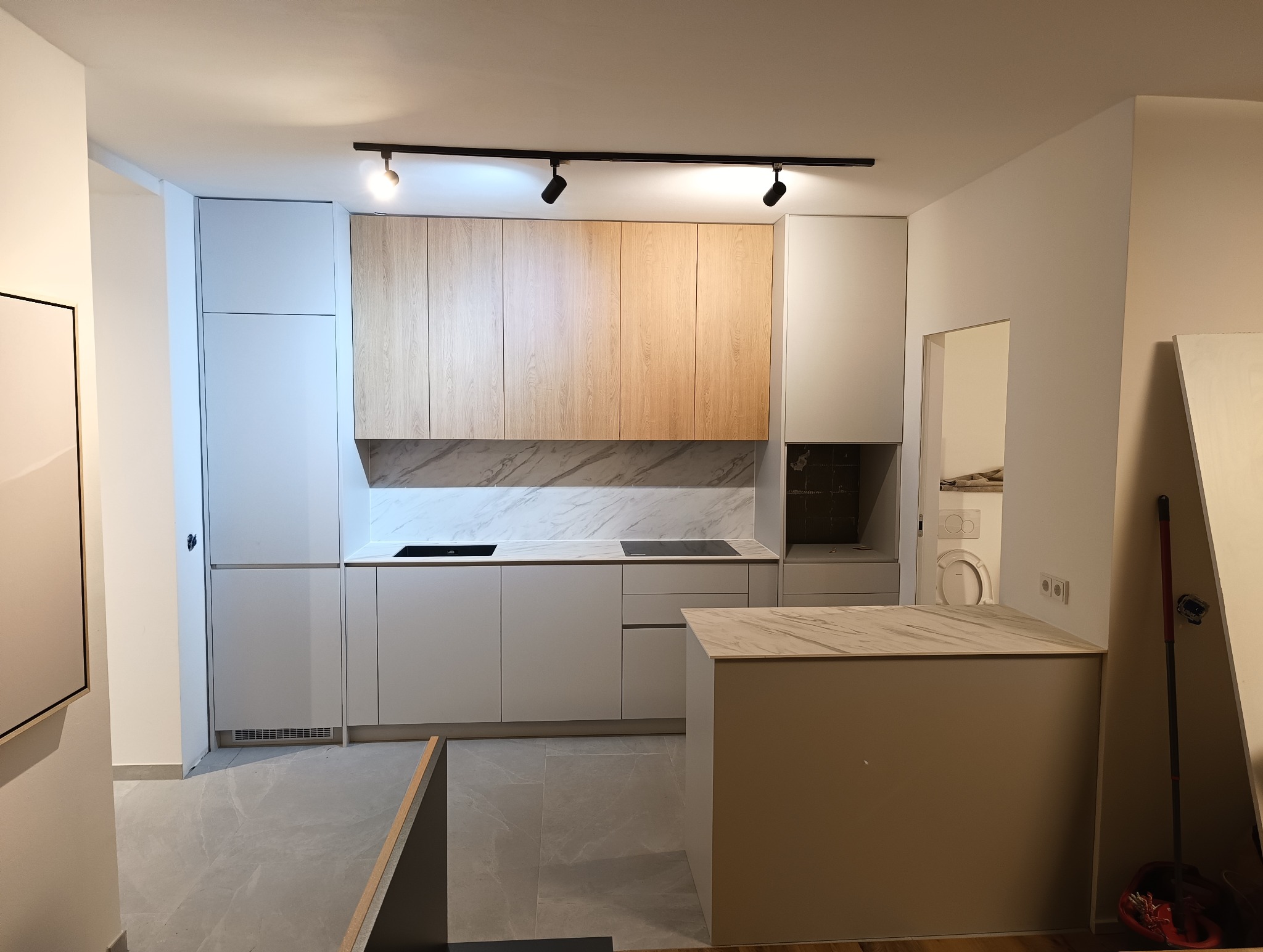 Nowoczesna kuchnia z wyspą, fronty w odcieniach bieli i drewna, marmurowy backsplash. Widoczne oświetlenie punktowe i fragment łazienki w tle.