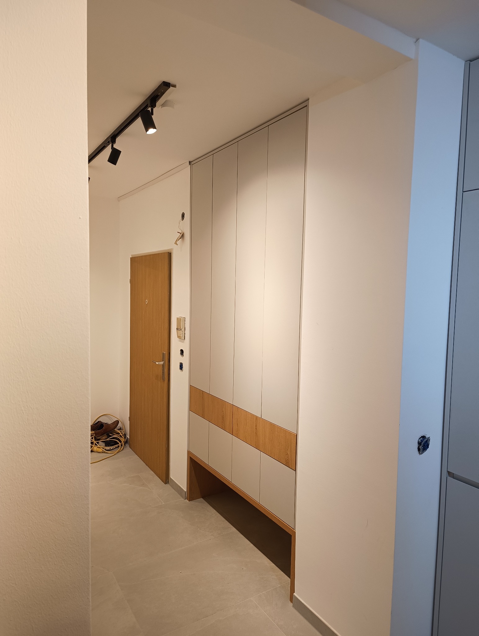 Nowoczesna szafa wnękowa z szarymi frontami i drewnianymi akcentami, wkomponowana w przedpokój z oświetleniem szynowym i drewnianymi drzwiami wejściowymi.