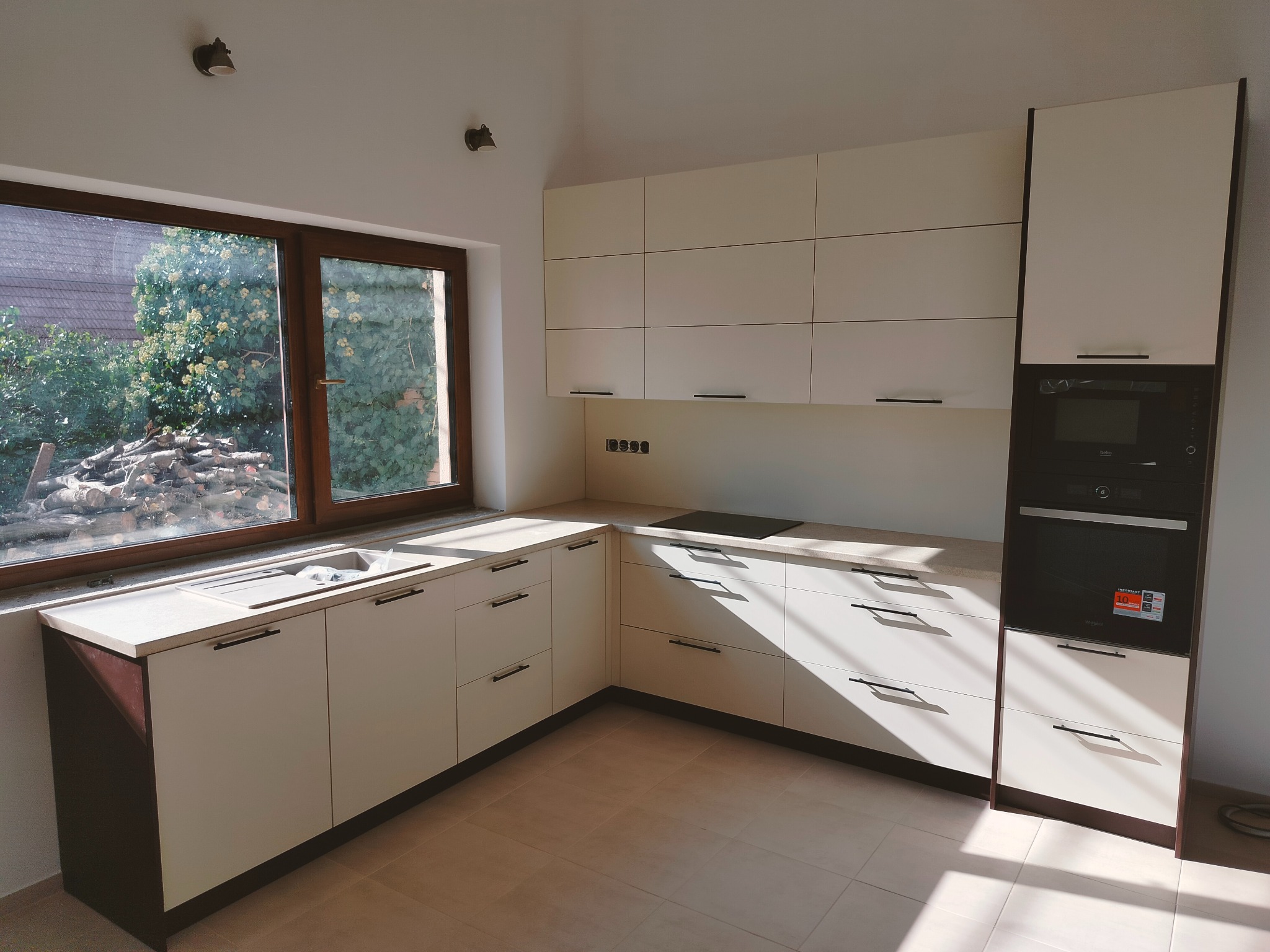 Jasna kuchnia z drewnianymi akcentami, widok na okno z stertą drewna na zewnątrz. Minimalistyczne uchwyty, nowoczesny piekarnik w zabudowie.