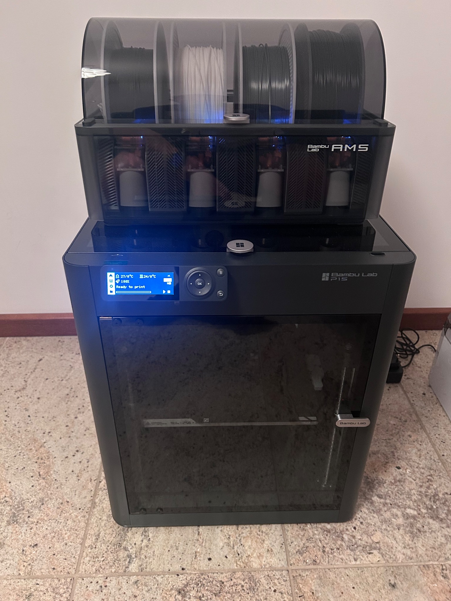 Czarna drukarka 3D Bambu Lab P1S z modułem AMS, widoczne szpule filamentu w różnych kolorach. Ekran drukarki podświetlony, gotowy do druku.