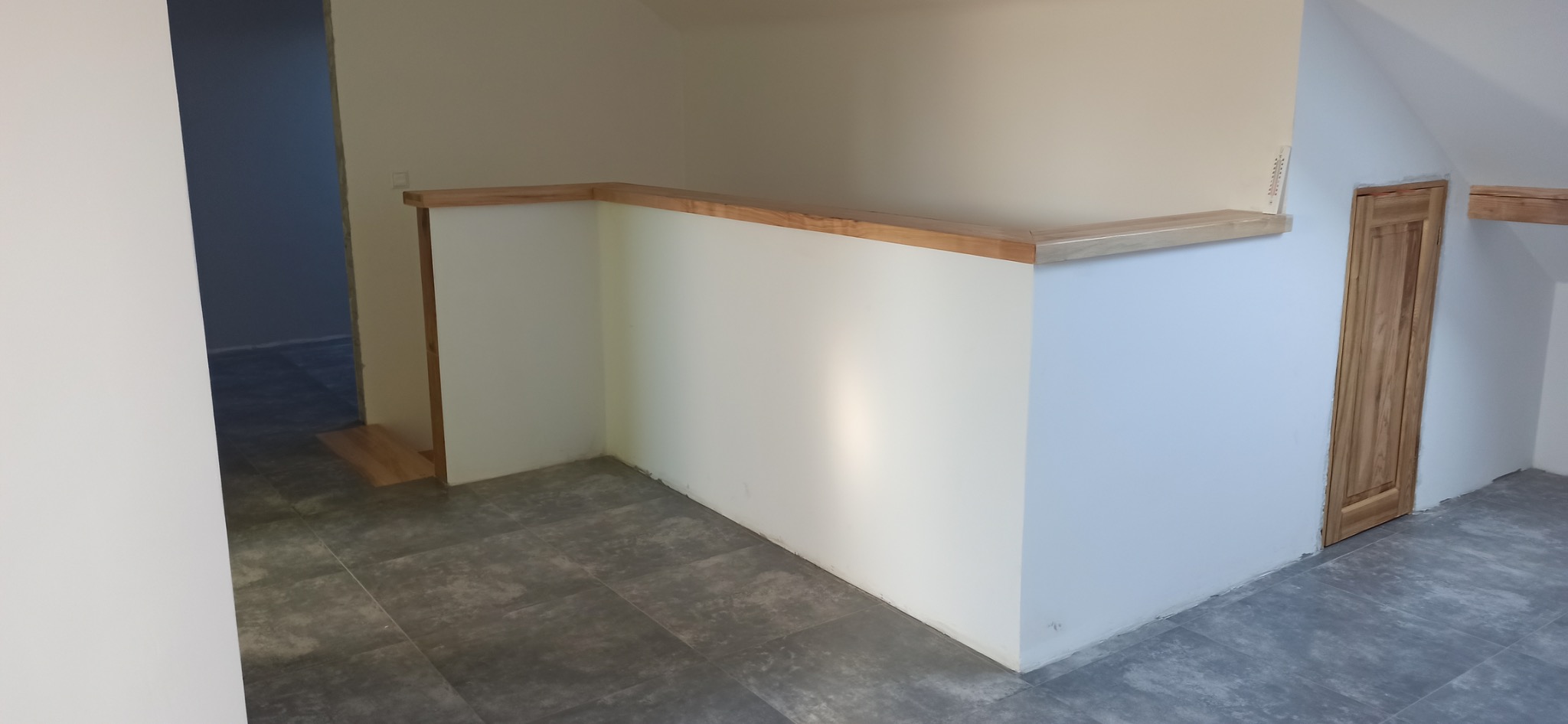 Wykończone poddasze z szarą podłogą, białą ścianką działową z drewnianym blatem i drewnianymi drzwiami. Widok na sąsiednie pomieszczenie.