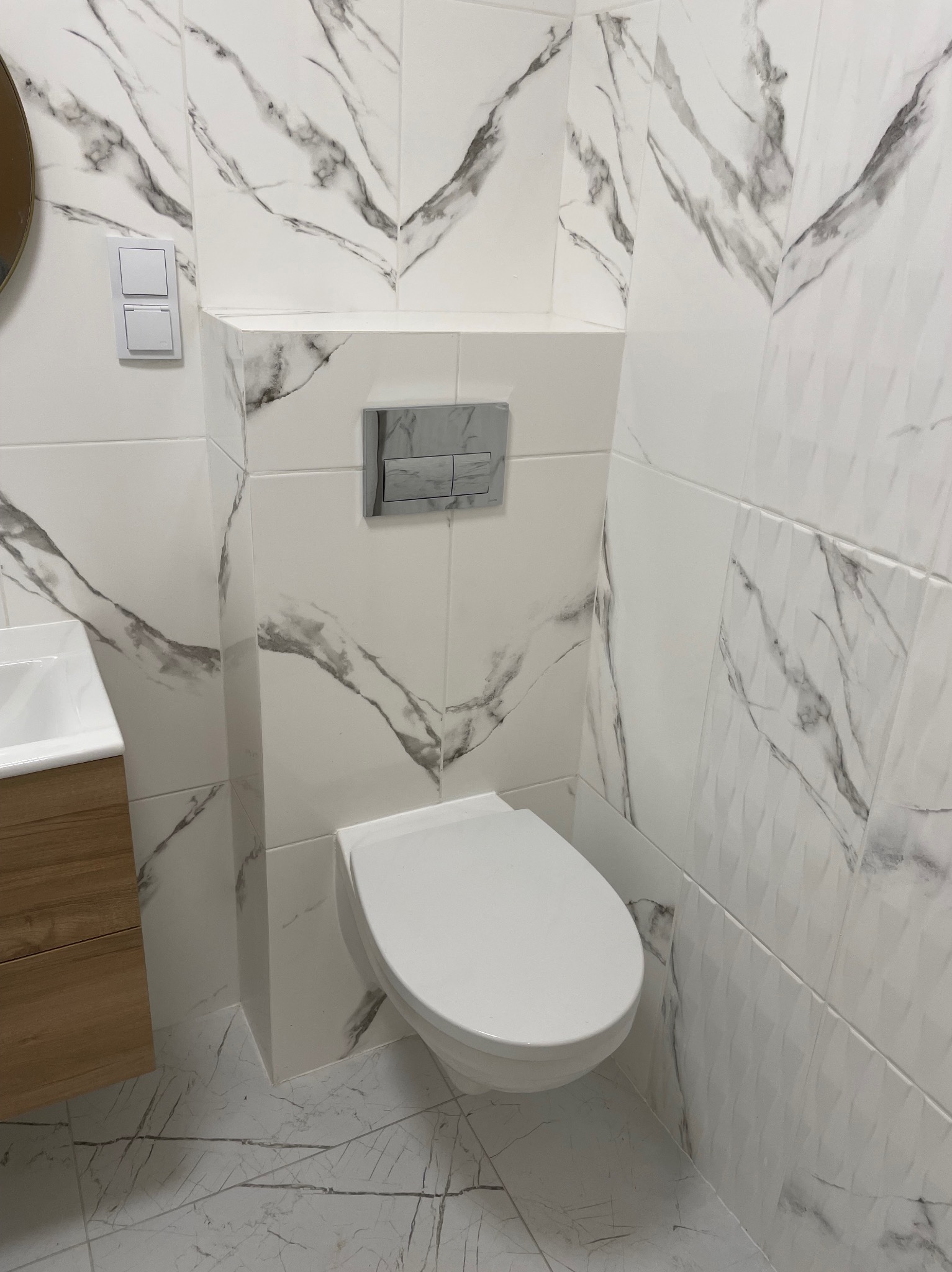 Biała toaleta z zabudową geberit i marmurowymi płytkami w łazience. Widoczny fragment szafki pod umywalką i włącznik światła. Nowoczesny design.