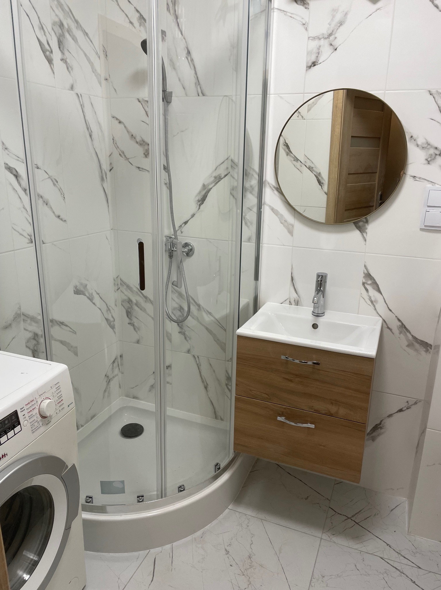 Nowoczesna łazienka z kabiną prysznicową, umywalką z szafką w odcieniach drewna i okrągłym lustrem. Białe płytki z szarym marmurkowym wzorem na ścianach i podłodze.