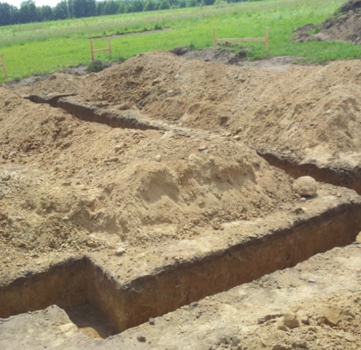 Wykopane fundamenty pod budowę domu na działce z zielonym trawnikiem w tle. Widoczne równe rowy w ziemi i pryzmy piasku.