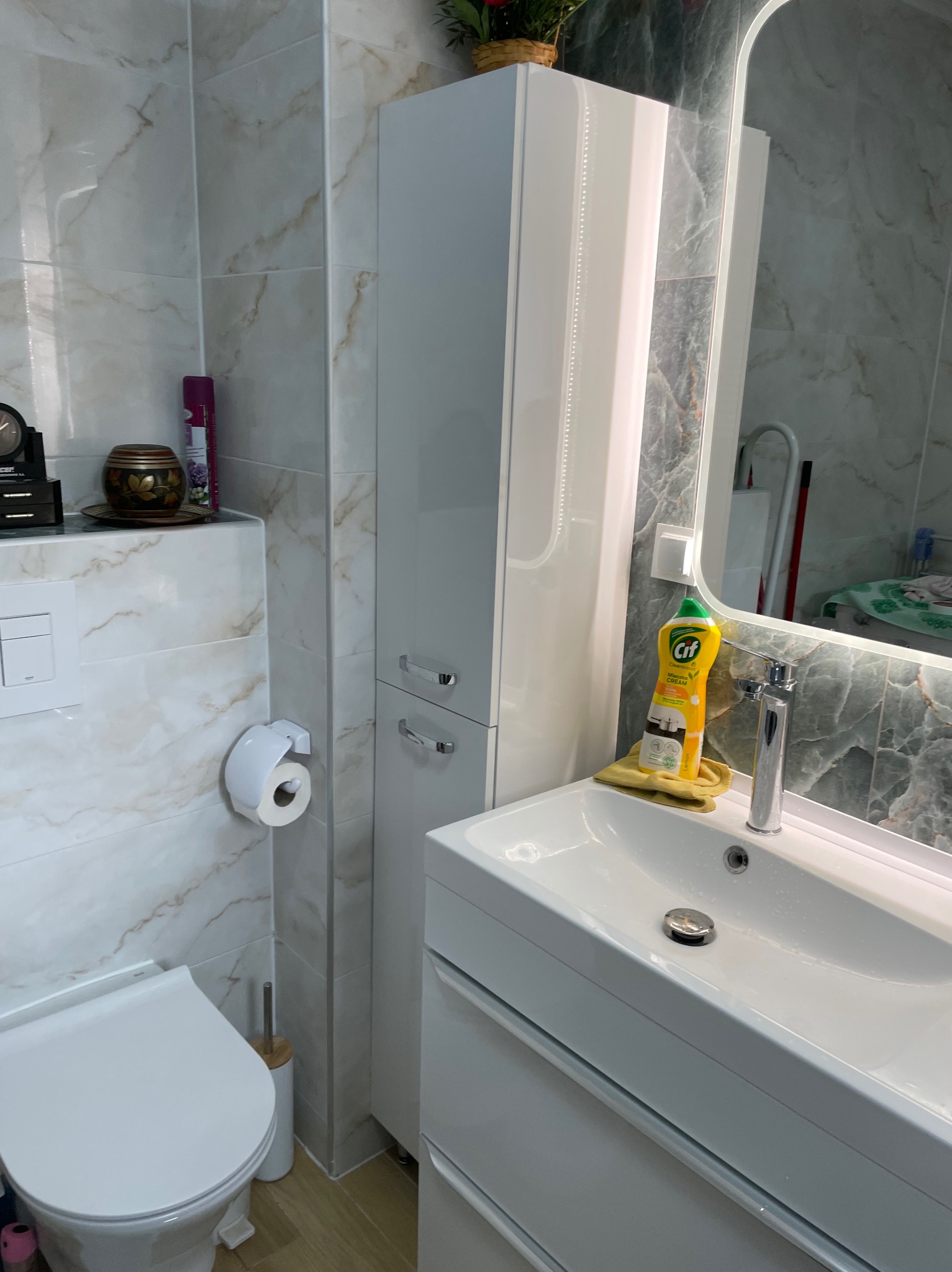 Nowoczesna łazienka z marmurowymi płytkami, białą szafką i umywalką. W tle lustro z podświetleniem LED. Widoczny fragment toalety i akcesoria łazienkowe.