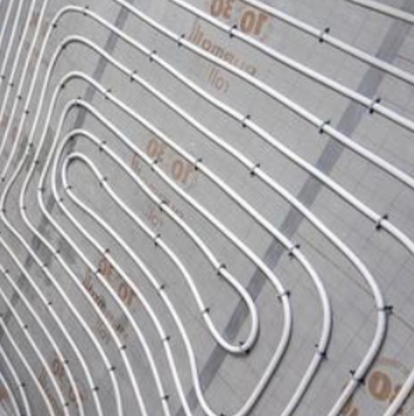 Białe rury ogrzewania podłogowego, równolegle ułożone na szarej macie izolacyjnej, przymocowane czarnymi uchwytami. Widok z góry, ukośny kadr.