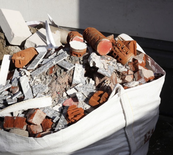 Worek z gruzem budowlanym: cegły, płytki, fragmenty betonu. Widoczne ślady zaprawy i różnorodność materiałów. Ujęcie z góry, ostre światło.