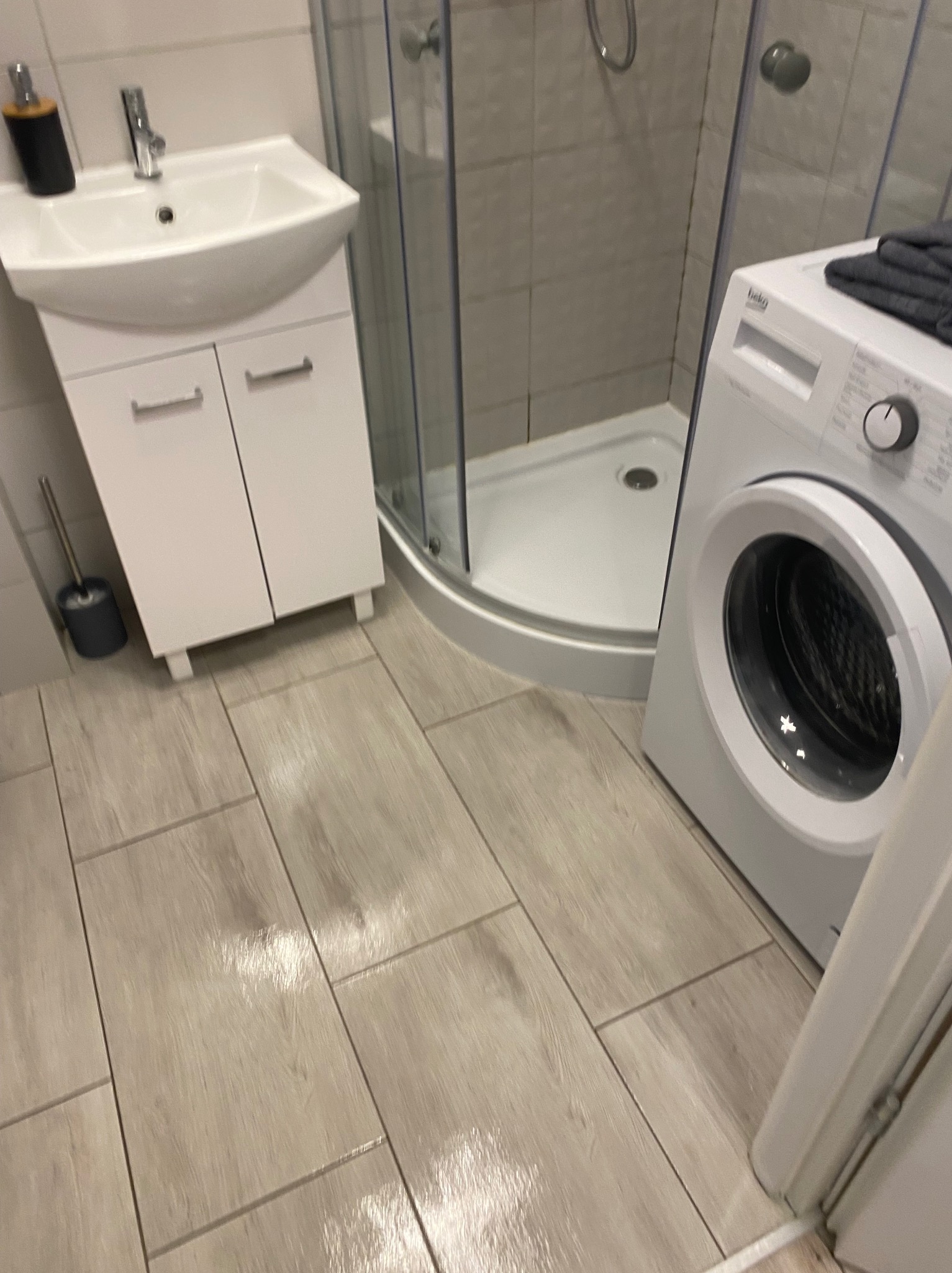 Jasna łazienka po remoncie: biała umywalka z szafką, kabina prysznicowa i pralka Beko. Podłoga wyłożona płytkami imitującymi drewno. Widok na fragment drzwi.