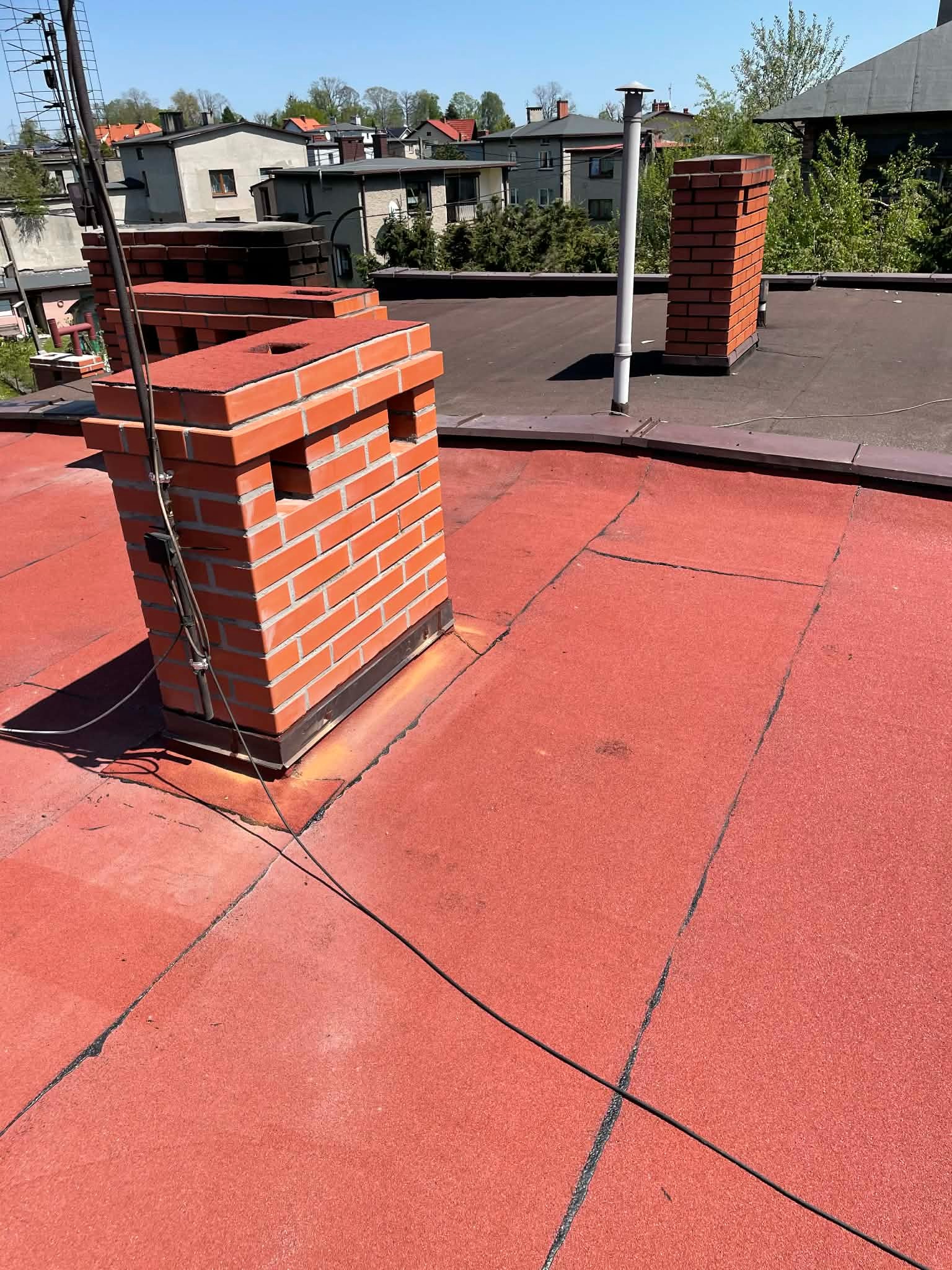 Czerwony dach pokryty papą z ceglanymi kominami w słoneczny dzień. Widoczne detale łączeń papy i instalacje na kominie. Tło stanowią budynki mieszkalne i drzewa.