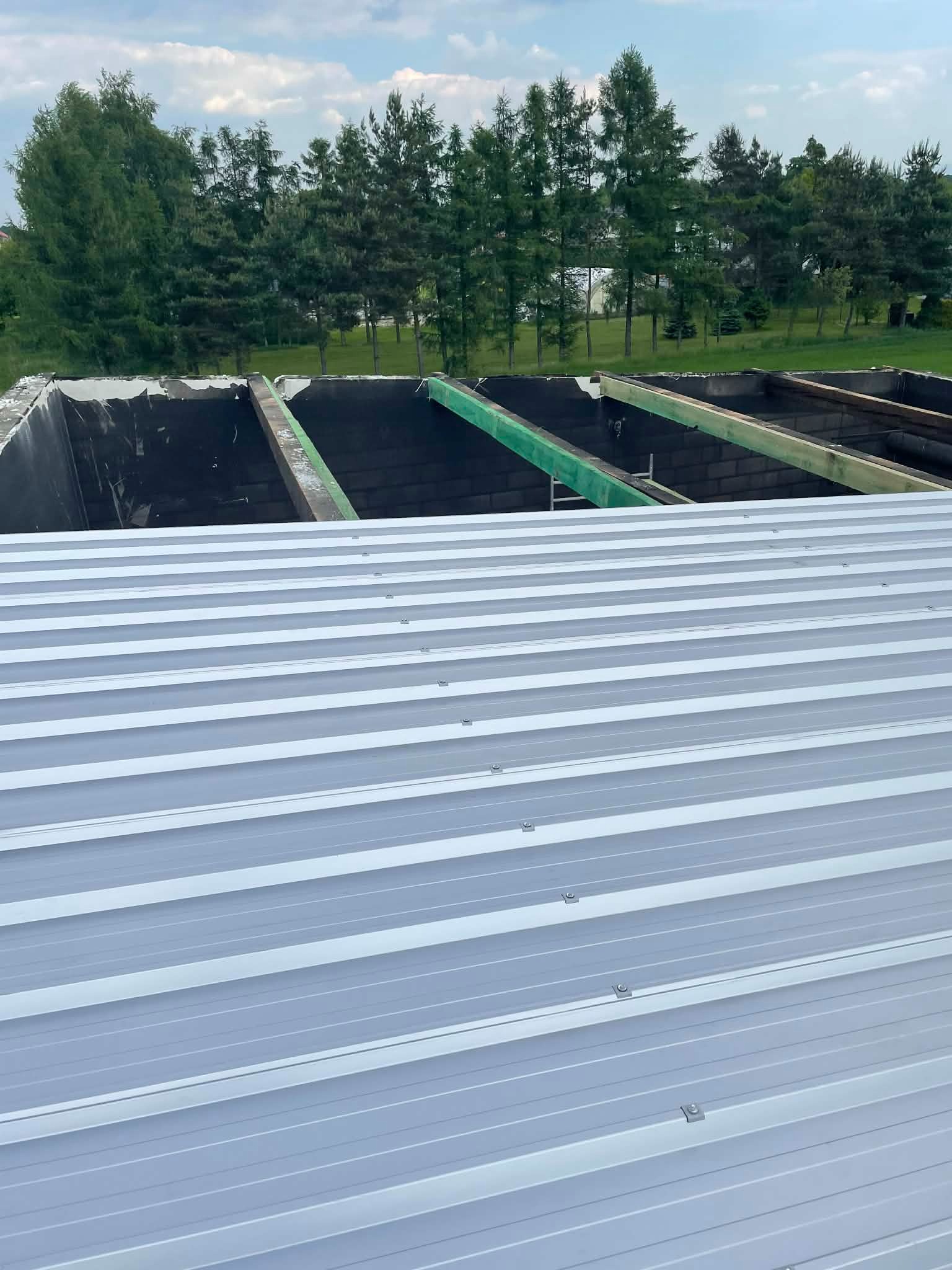 Szary blaszany dach w trakcie montażu, widoczne drewniane elementy konstrukcyjne i tło z zielenią drzew. Dach z blachy trapezowej, mocowany na wkręty.