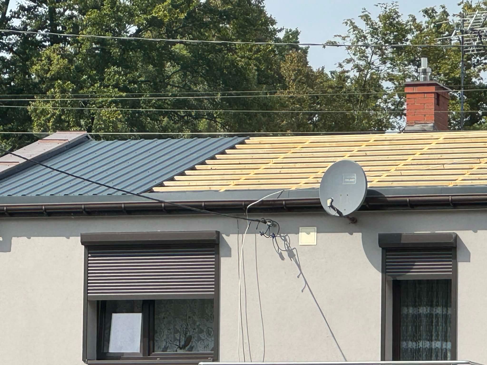 Częściowo wymieniony dach z blachy trapezowej, widoczne łaty, komin z cegły, antena satelitarna na elewacji budynku z brązowymi roletami okiennymi.