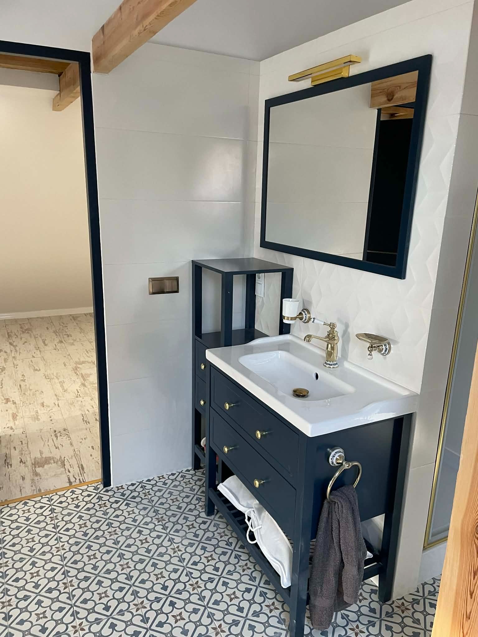 Wykończona łazienka z granatową szafką pod umywalką, złotą armaturą i wzorzystą podłogą. Widoczne detale: lustro w granatowej ramie, drewniana belka stropowa.