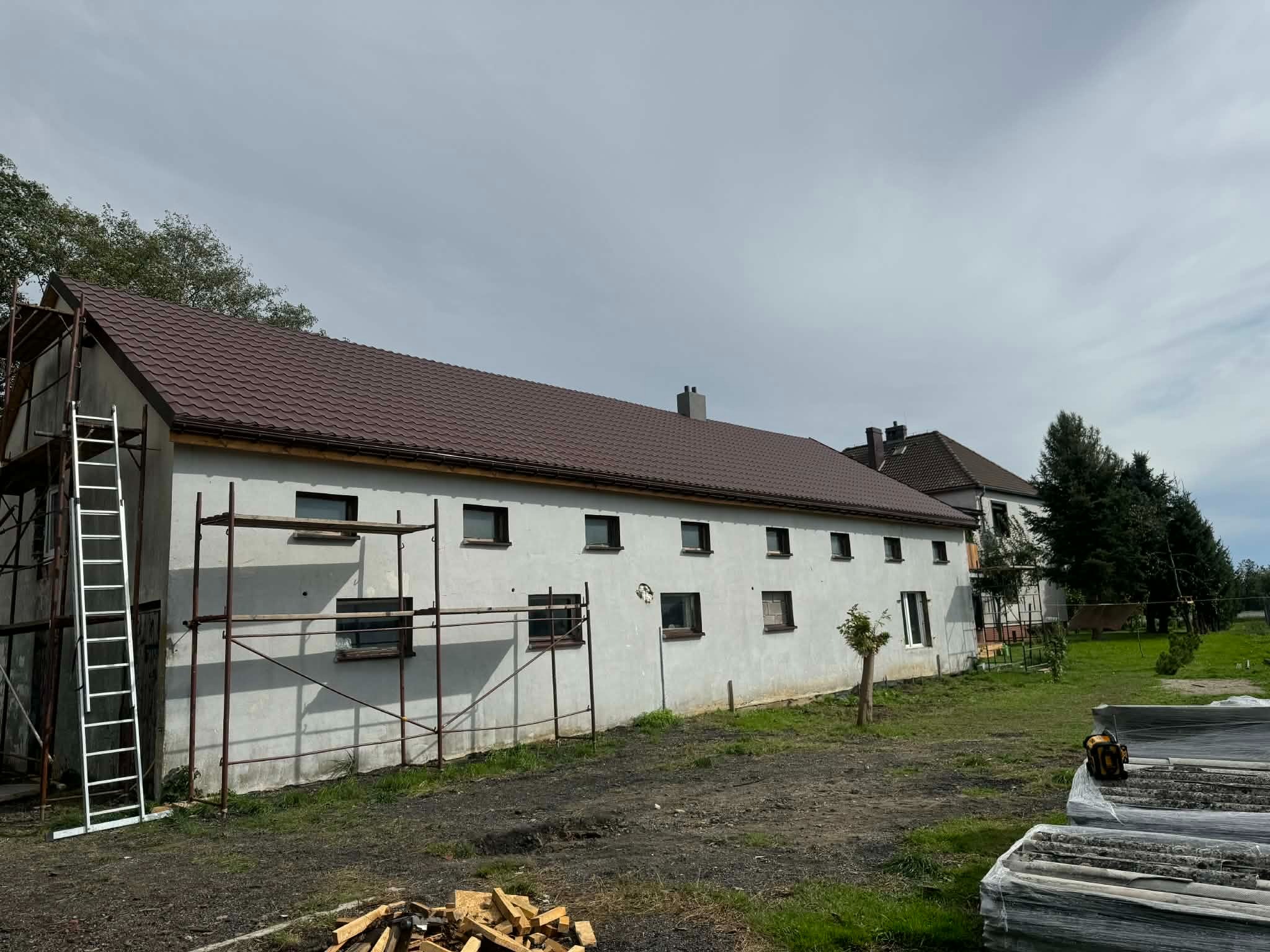 Budynek w trakcie remontu elewacji i wymiany dachu. Widoczne rusztowania, nowa, brązowa dachówka i przygotowane materiały budowlane. Prace w toku.