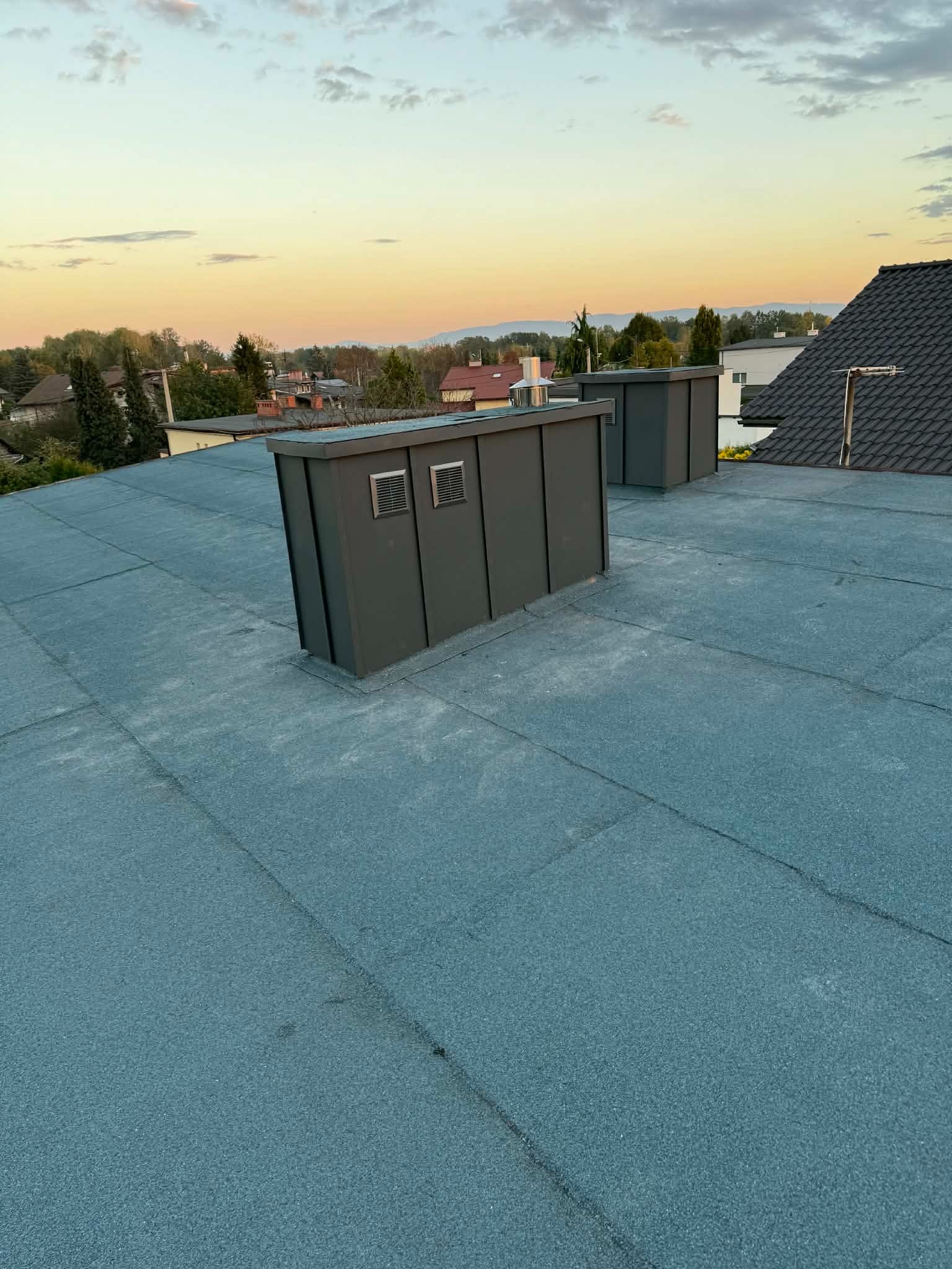 Płaski dach pokryty papą termozgrzewalną z dwoma szarymi kominami wentylacyjnymi. W tle zabudowa jednorodzinna i zachodzące słońce.
