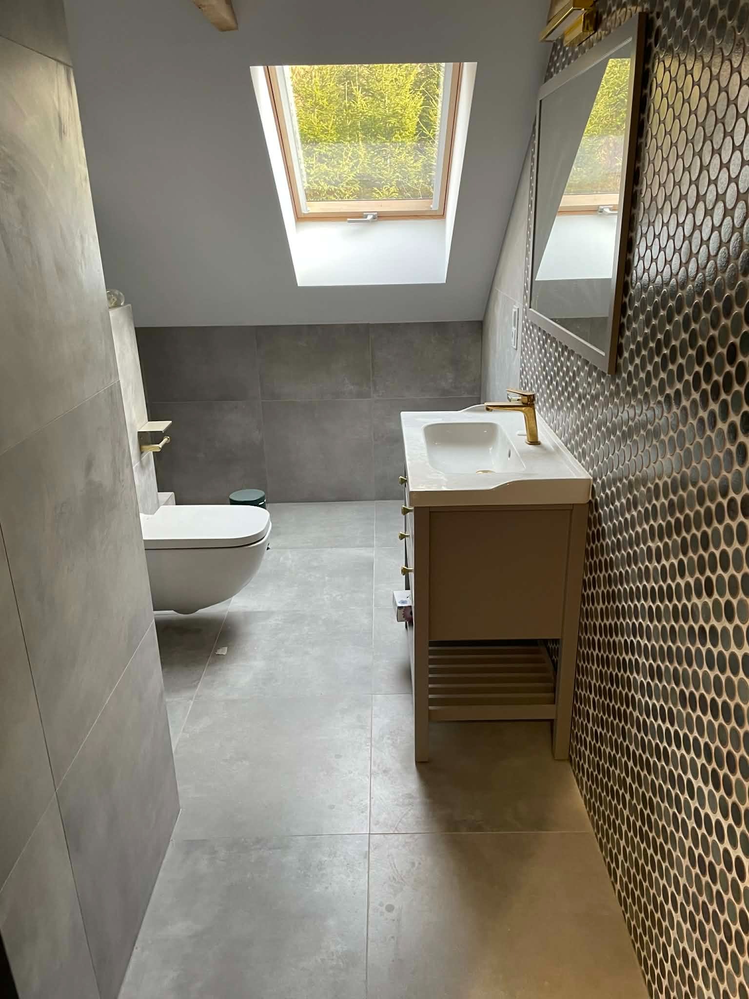Nowoczesna łazienka na poddaszu z szarą glazurą, umywalką z szafką i lustrem. Okno dachowe wpuszcza naturalne światło. Minimalistyczny design z akcentami złota.