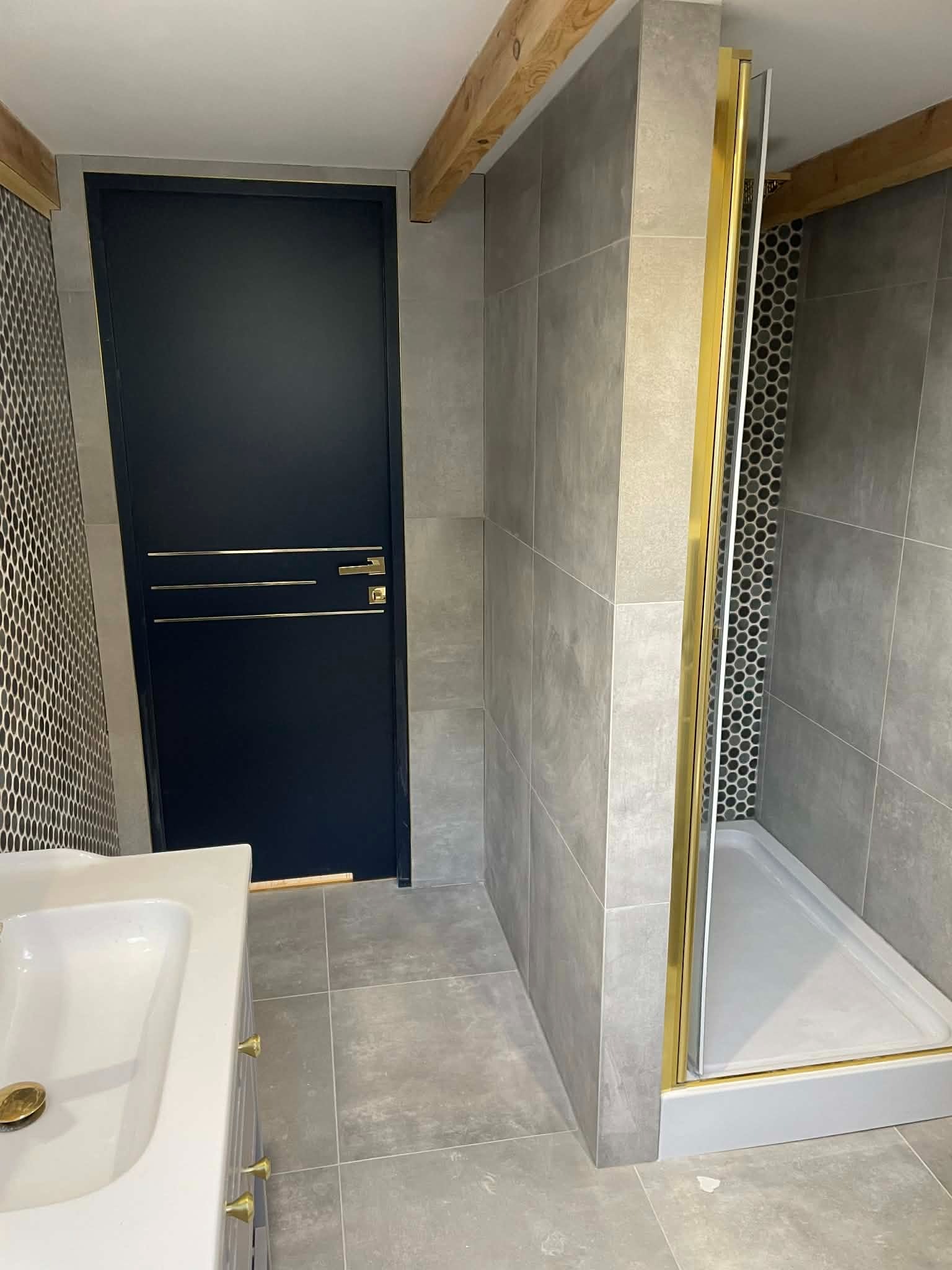 Wykończona łazienka z kabiną prysznicową w złotych ramach, umywalką i ciemnymi drzwiami z złotymi akcentami. Szare płytki i drewniane belki dodają elegancji.