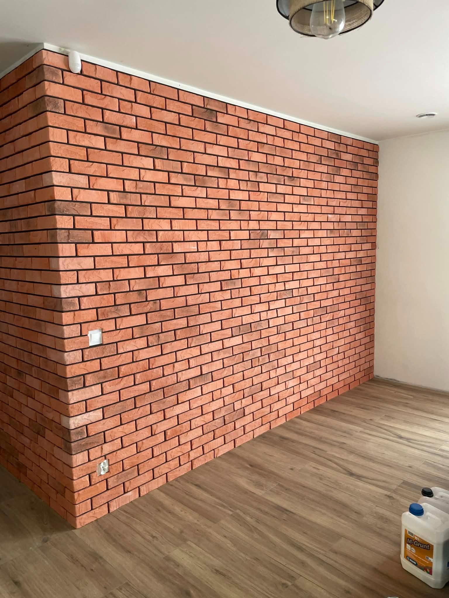 Ściana wykończona czerwoną cegłą klinkierową w nowocześnie urządzonym wnętrzu z drewnianą podłogą i widocznymi puszkami elektrycznymi. Widoczna lampa sufitowa.