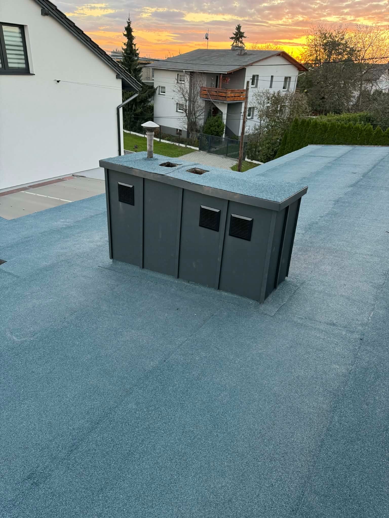 Niebieski dach pokryty papą z szarą obudową kominową na tle domów i zachodzącego słońca. Widoczne detale wykończenia i faktura papy.