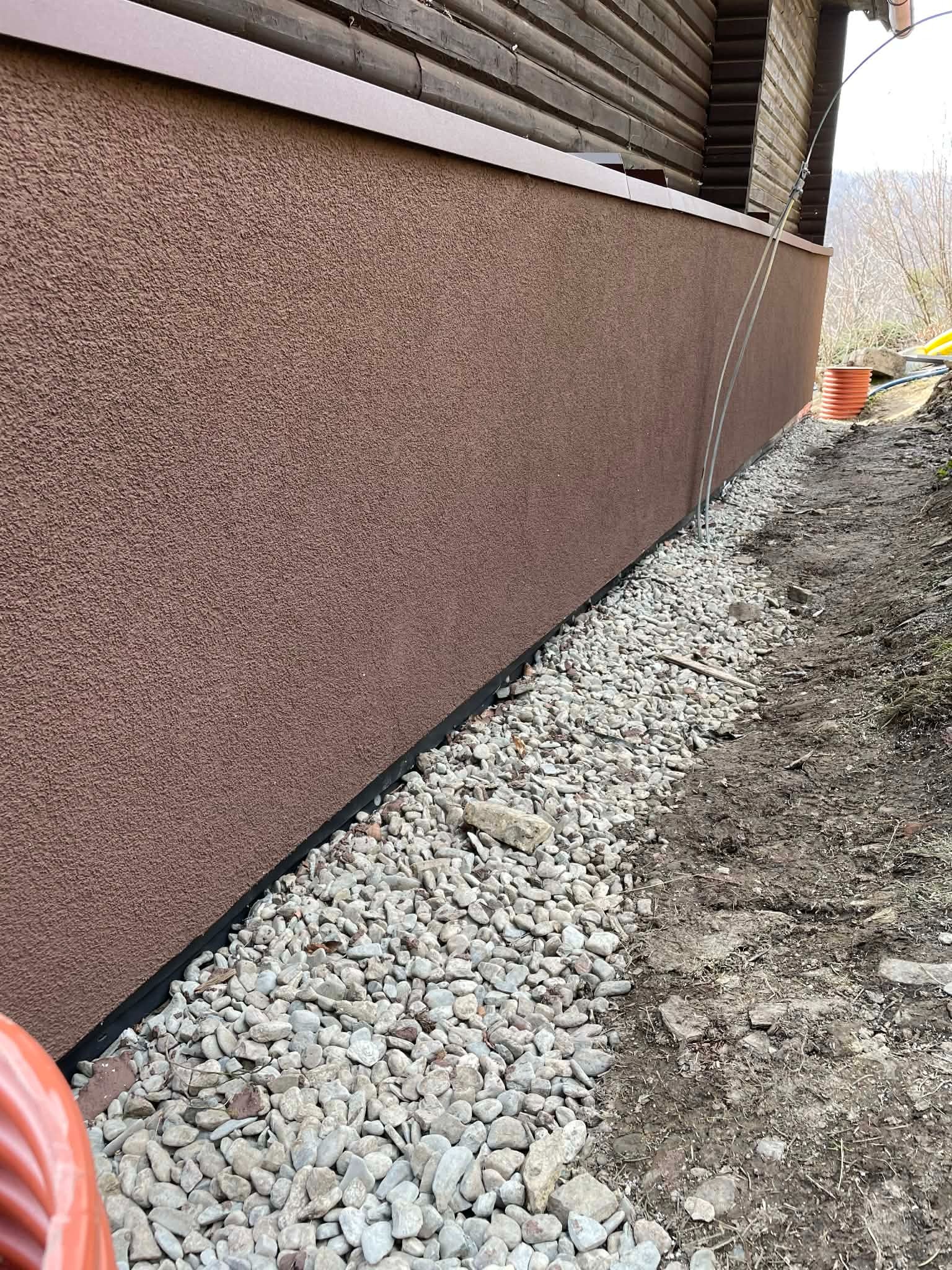 Brązowa elewacja domu z widoczną warstwą izolacyjną i drenażem z białych kamieni. Ujęcie z poziomu gruntu, podkreślające detale wykończenia.