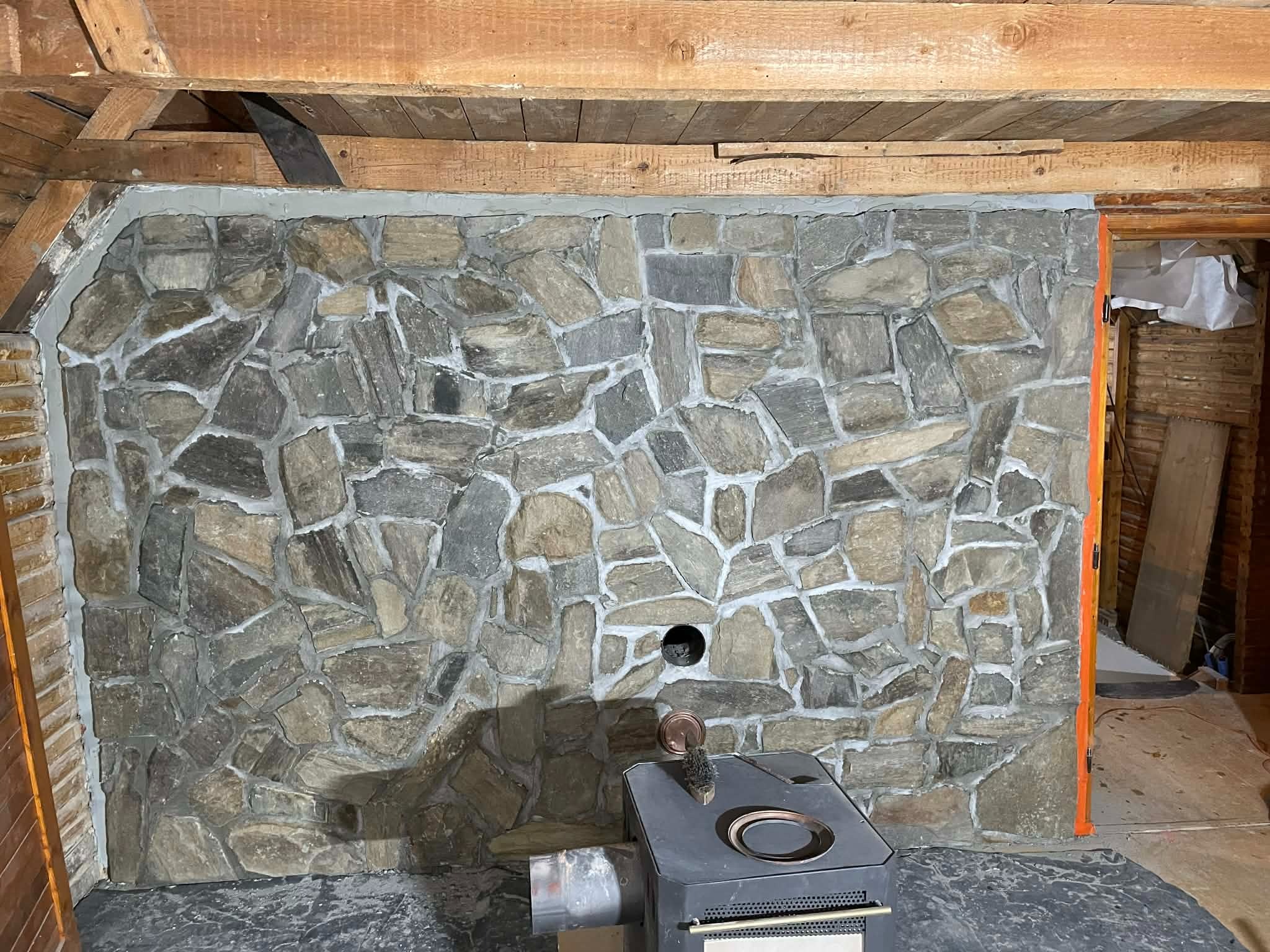 Ściana wyłożona nieregularnym kamieniem w odcieniach szarości i beżu, z widocznym kominem i drewnianą konstrukcją stropu. Fragment pieca na pierwszym planie.