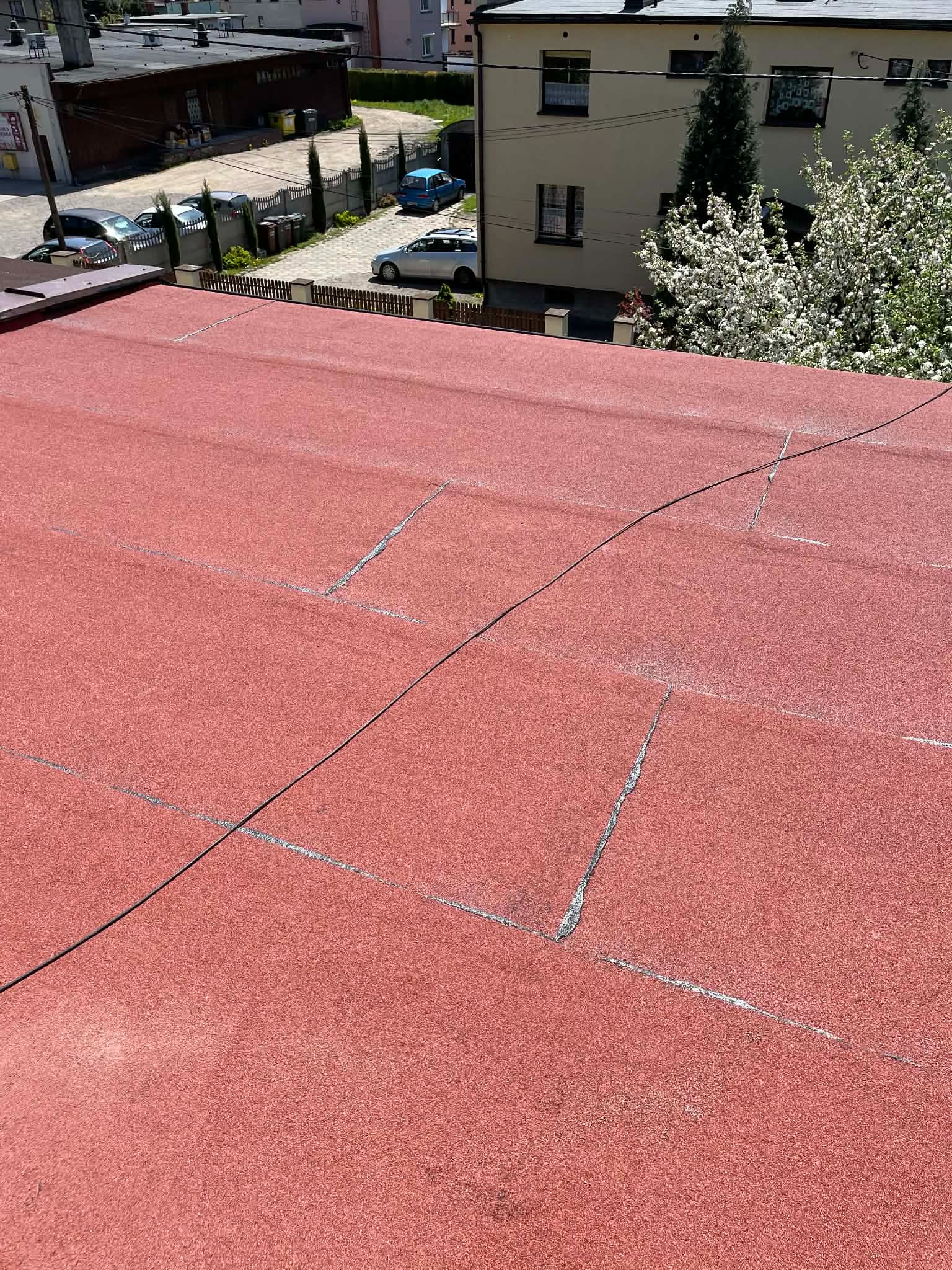 Czerwony dach pokryty papą termozgrzewalną z widocznymi łączeniami i przewodami, na tle zabudowań i zieleni. Widok z góry.