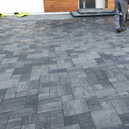Roman Luhovyj - Szara kostka brukowa ułożona na podjeździe, widoczne ubrania robocze i fragment osoby. Nowoczesny wzór kostki, świeżo po ułożeniu, zarys domu w tle.