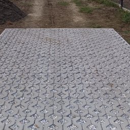 Roman Luhovyj - Ułożona szara ażurowa kostka brukowa wypełniona drobnym kamieniem na podwórku, widok z góry, w tle przyczepka i hałda ziemi.